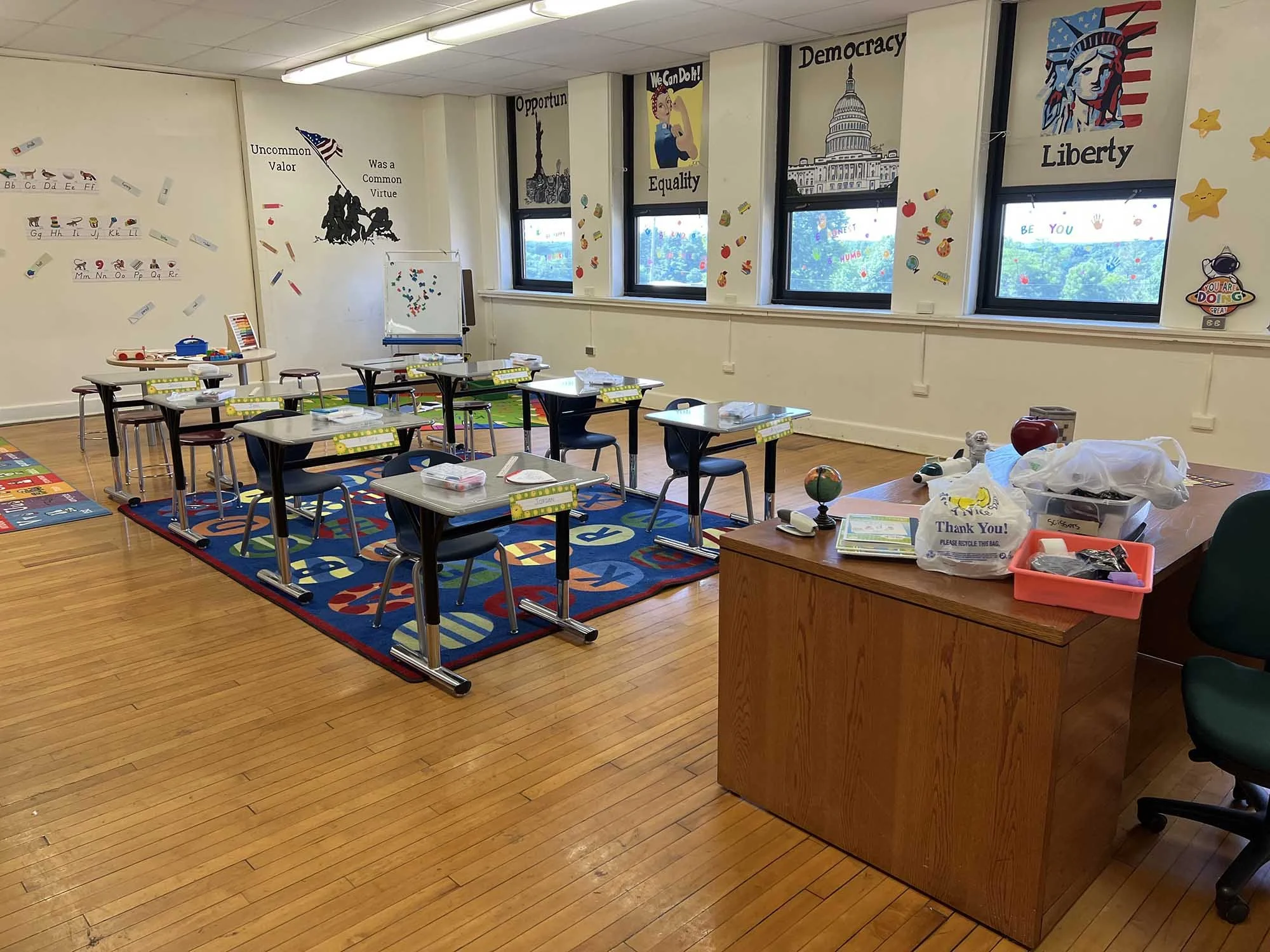 A classroom with desks, chairs, and educational decorations, including posters with themes of democracy, liberty, equality, and opportunity. Windows let in natural light, and the room has a colorful rug and some scattered supplies.
