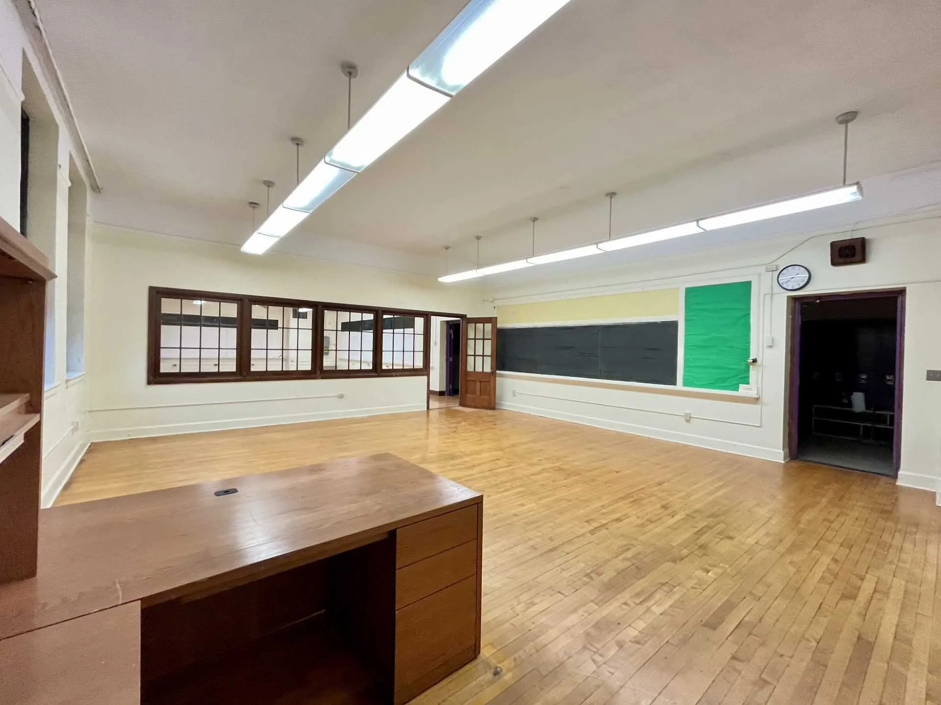 Empty classroom or meeting room with wooden floors, large windows, chalkboard, and fluorescent ceiling lights.