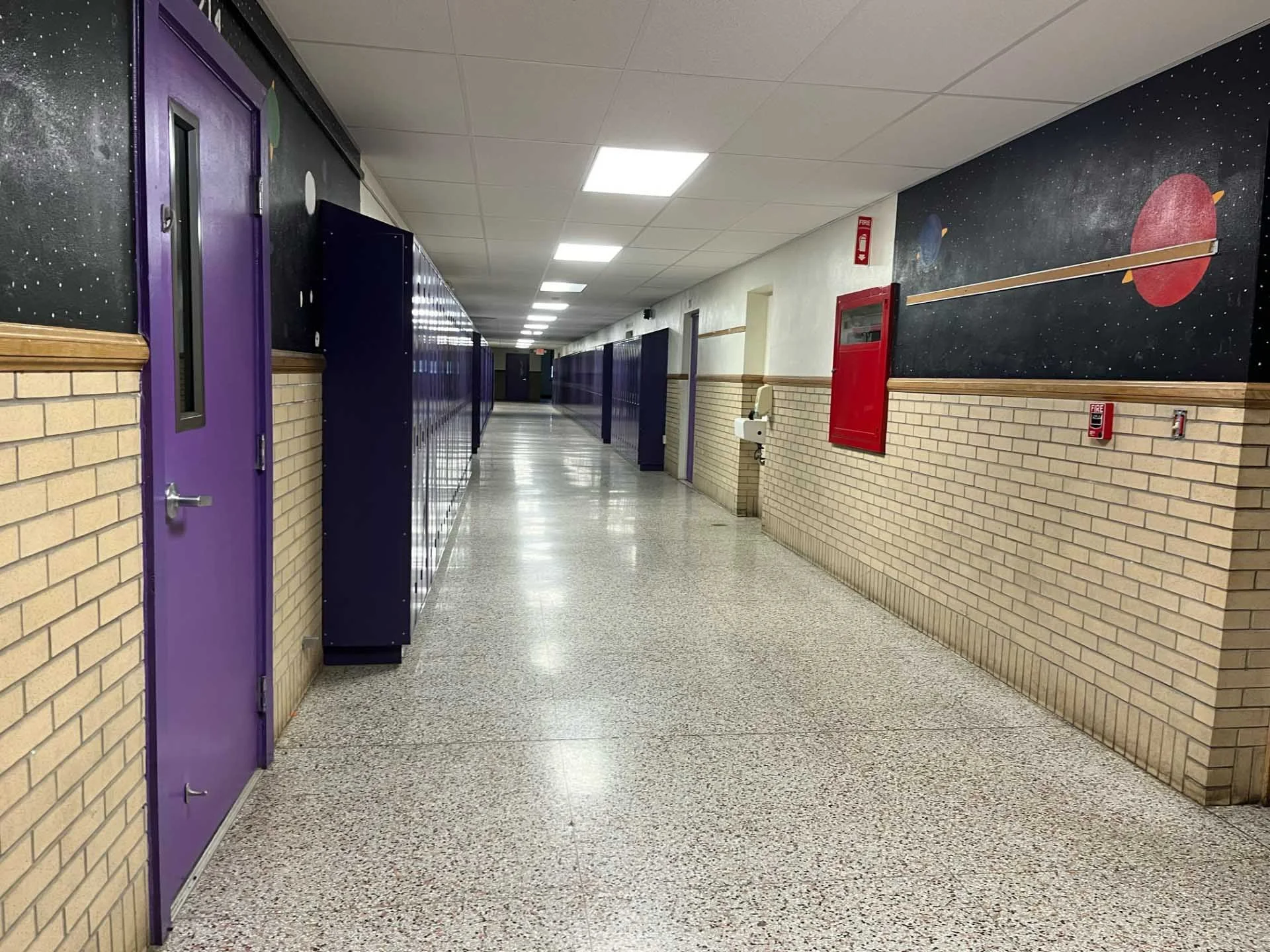 A school hallway with lockers, purple doors, and brick walls decorated with space-themed murals including planets and stars.