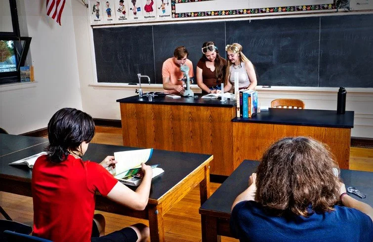 Students conducting a science experiment in a classroom with a blackboard and science posters, while other students observe.