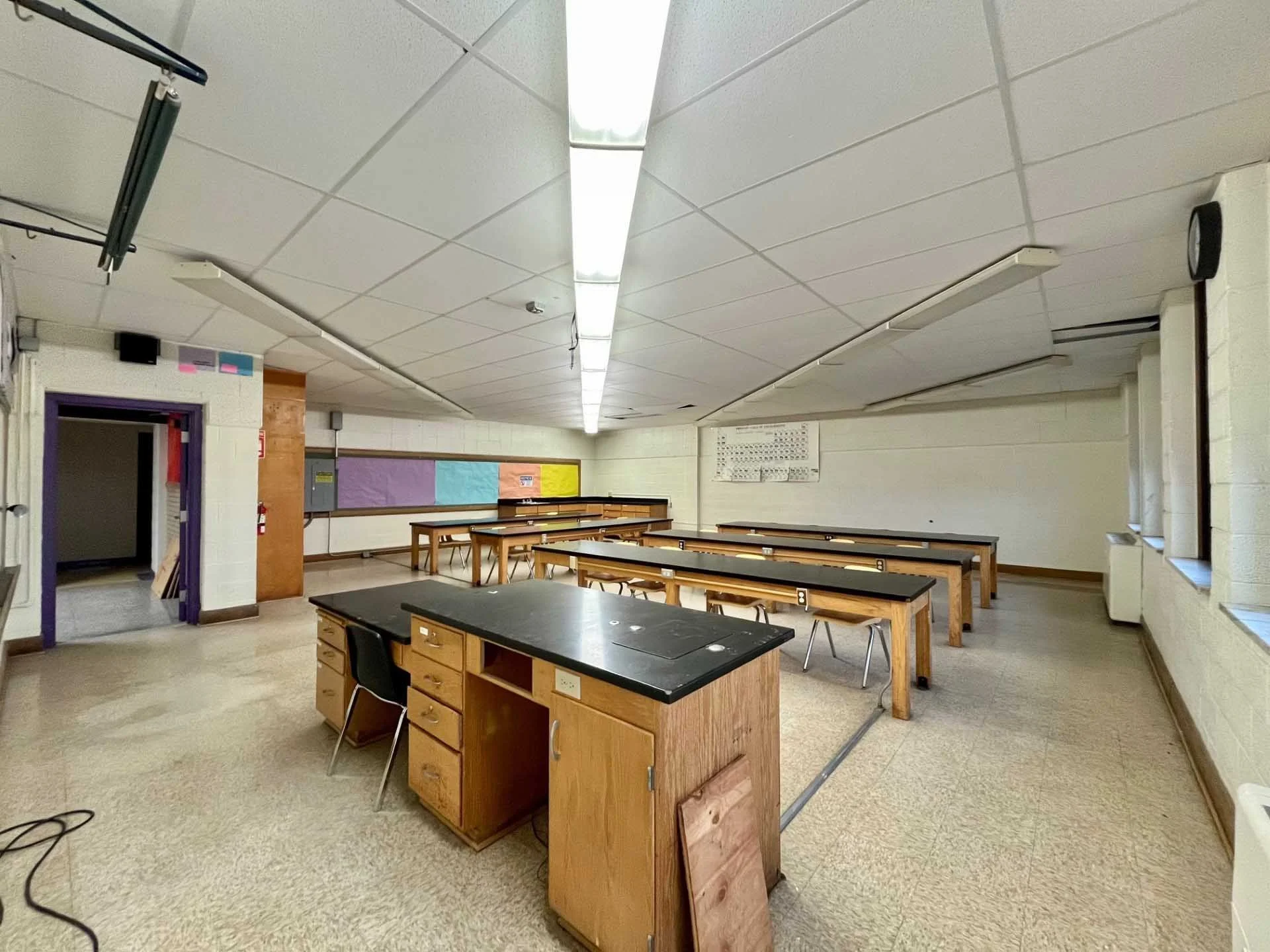 Empty classroom with wooden desks and black countertops, fluorescent ceiling lights, a purple door, and colorful bulletin boards on the wall.
