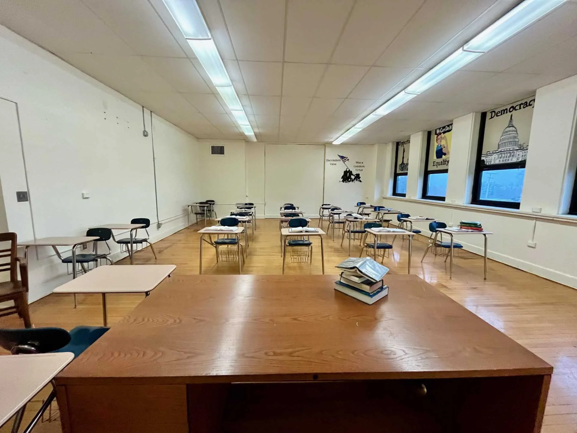 Empty classroom with desks and chairs, some stacked books on a desk, windows with posters, and a wall mural with patriotic and political messages.