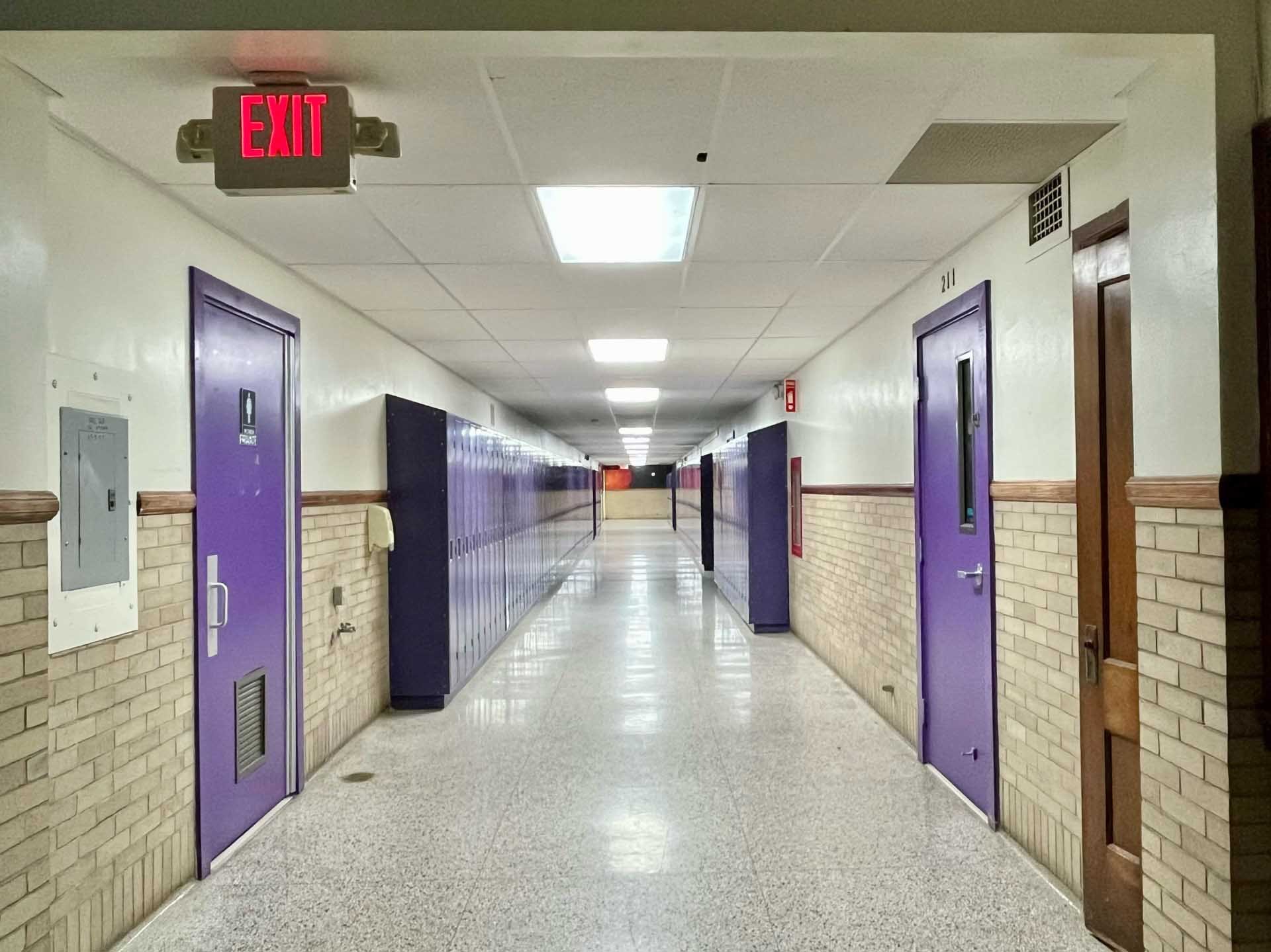 Hallway with purple doors, lockers, and an exit sign on the ceiling.