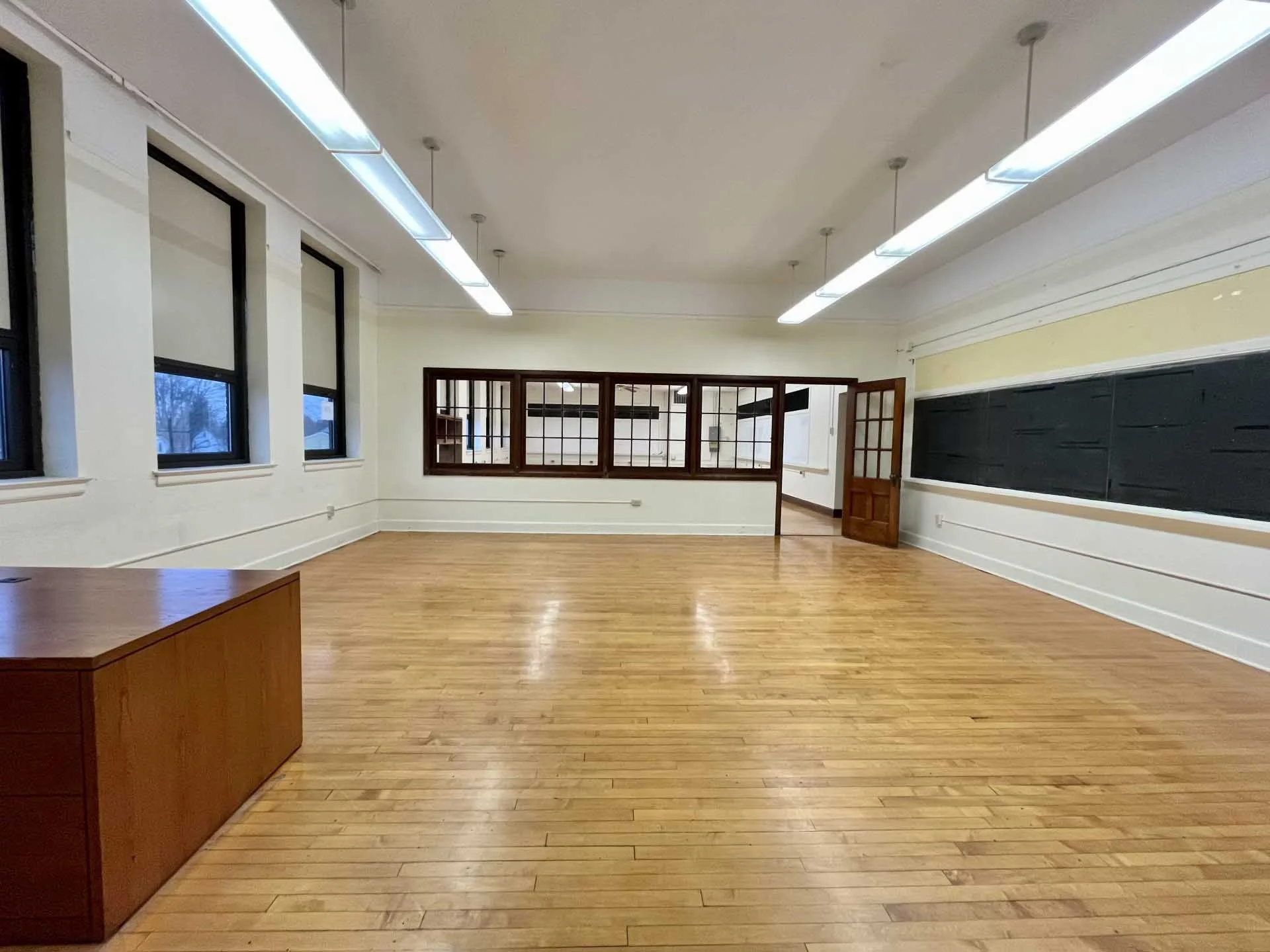 Empty classroom with wooden floors, large windows, a blackboard on the wall, fluorescent lights on the ceiling, and a wooden door.