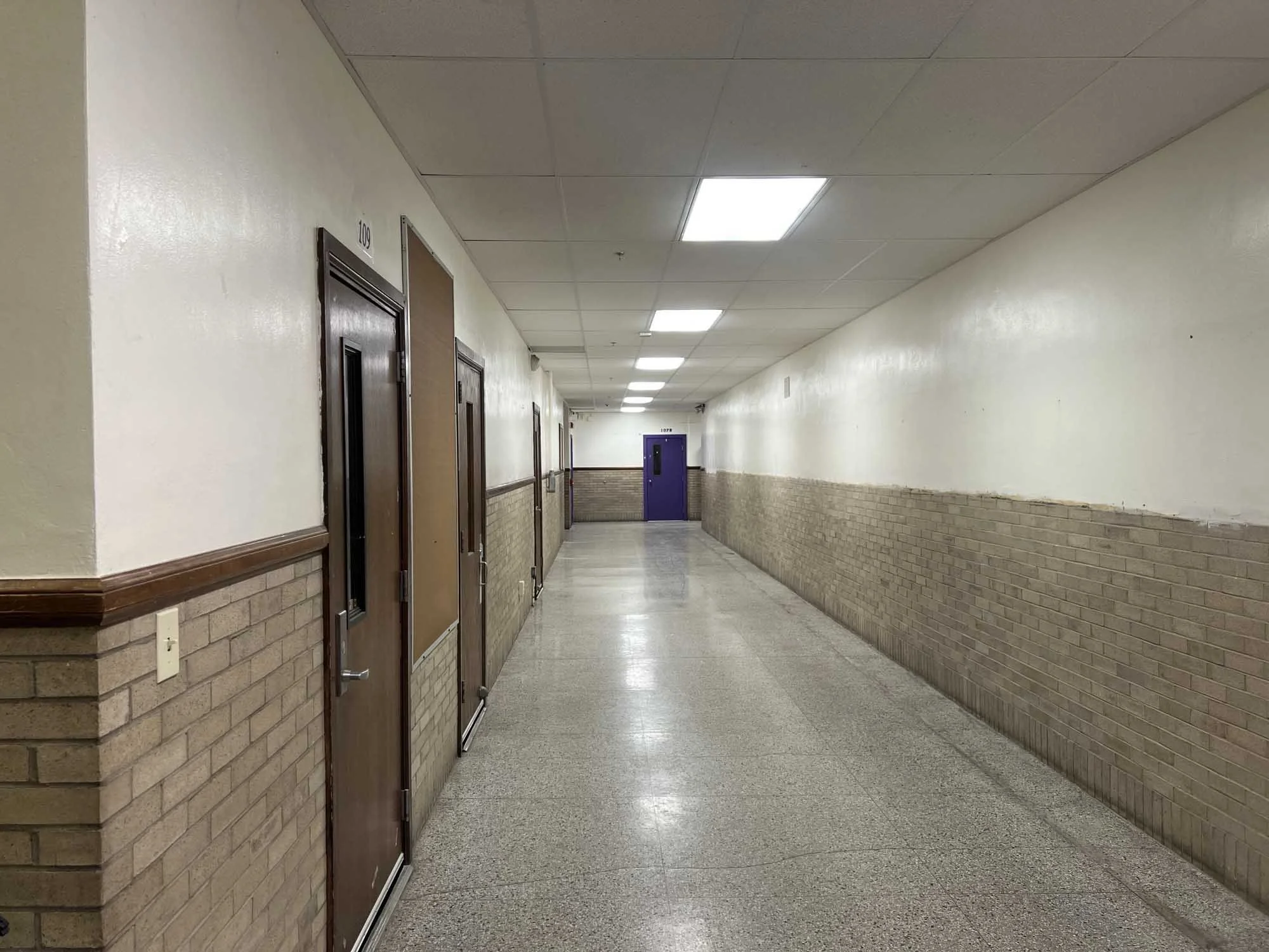 Hallway with wood and brick walls, fluorescent ceiling lights, and purple door at the end.