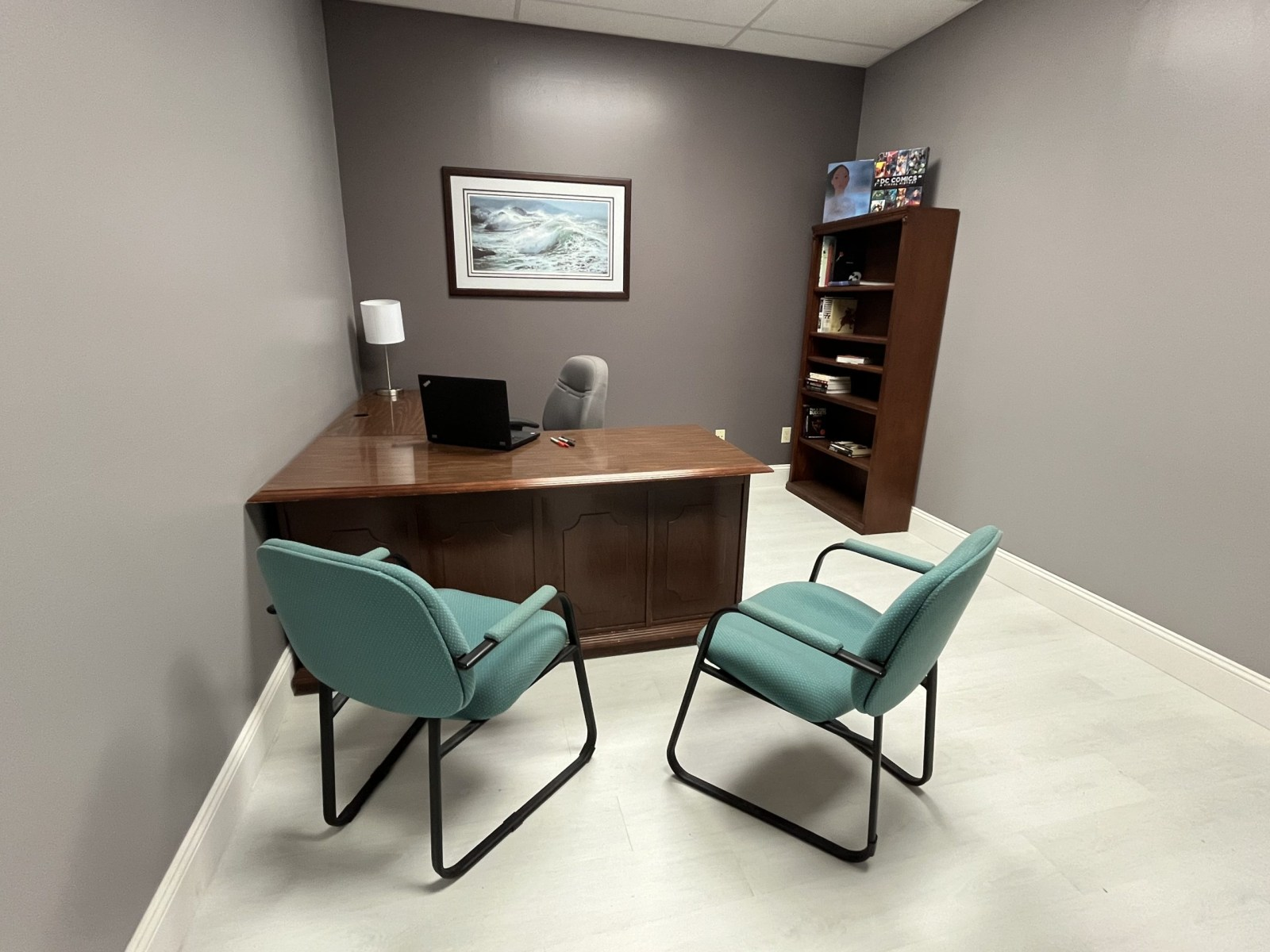 An office with a wooden desk, two teal chairs, a bookshelf, a framed wave painting on a gray wall, a laptop, a table lamp, and a few books and items on the shelf.
