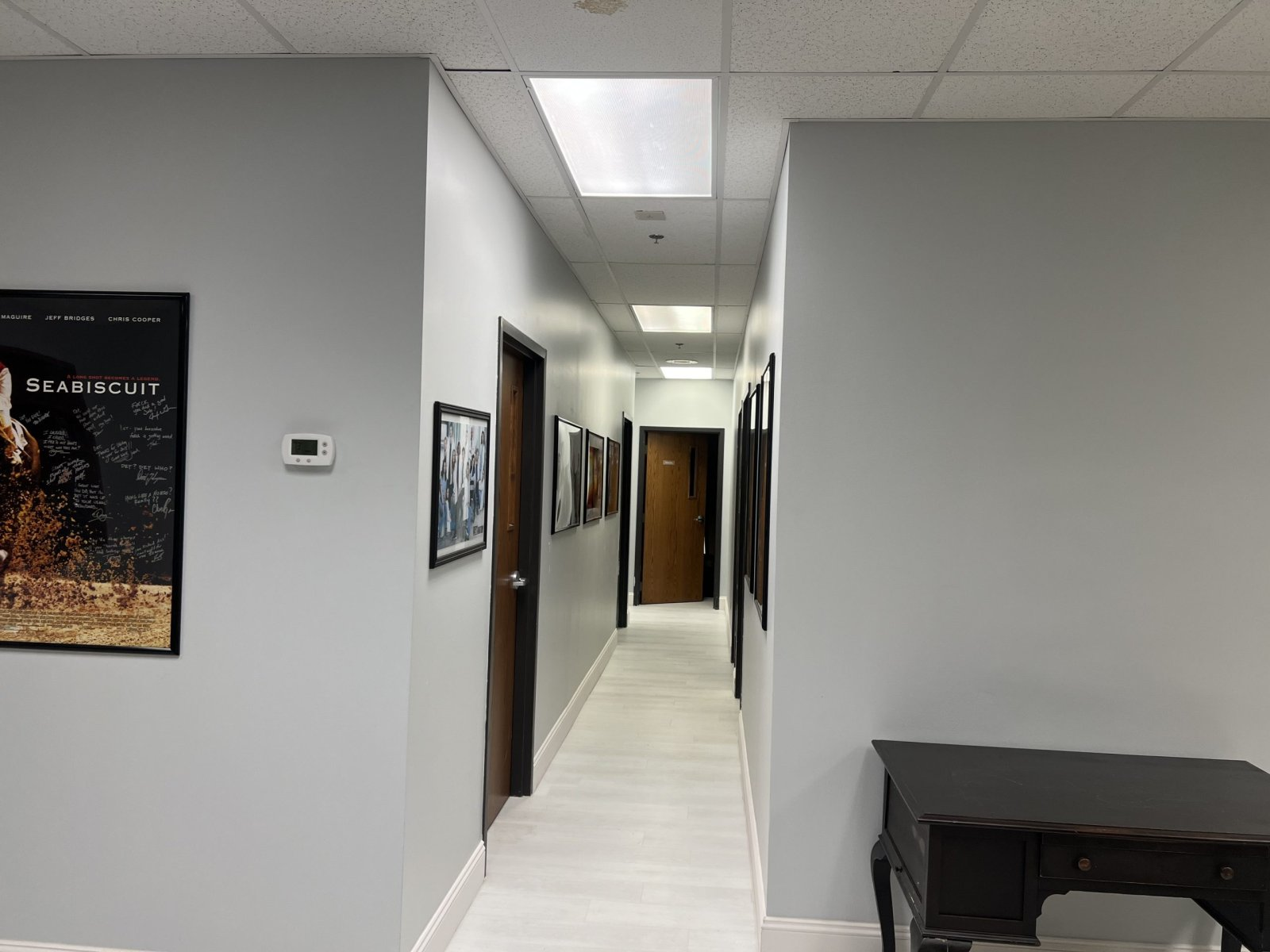 Empty office hallway with light gray walls, related framed pictures, closed wooden doors, ceiling lights, and a small dark desk on the right.