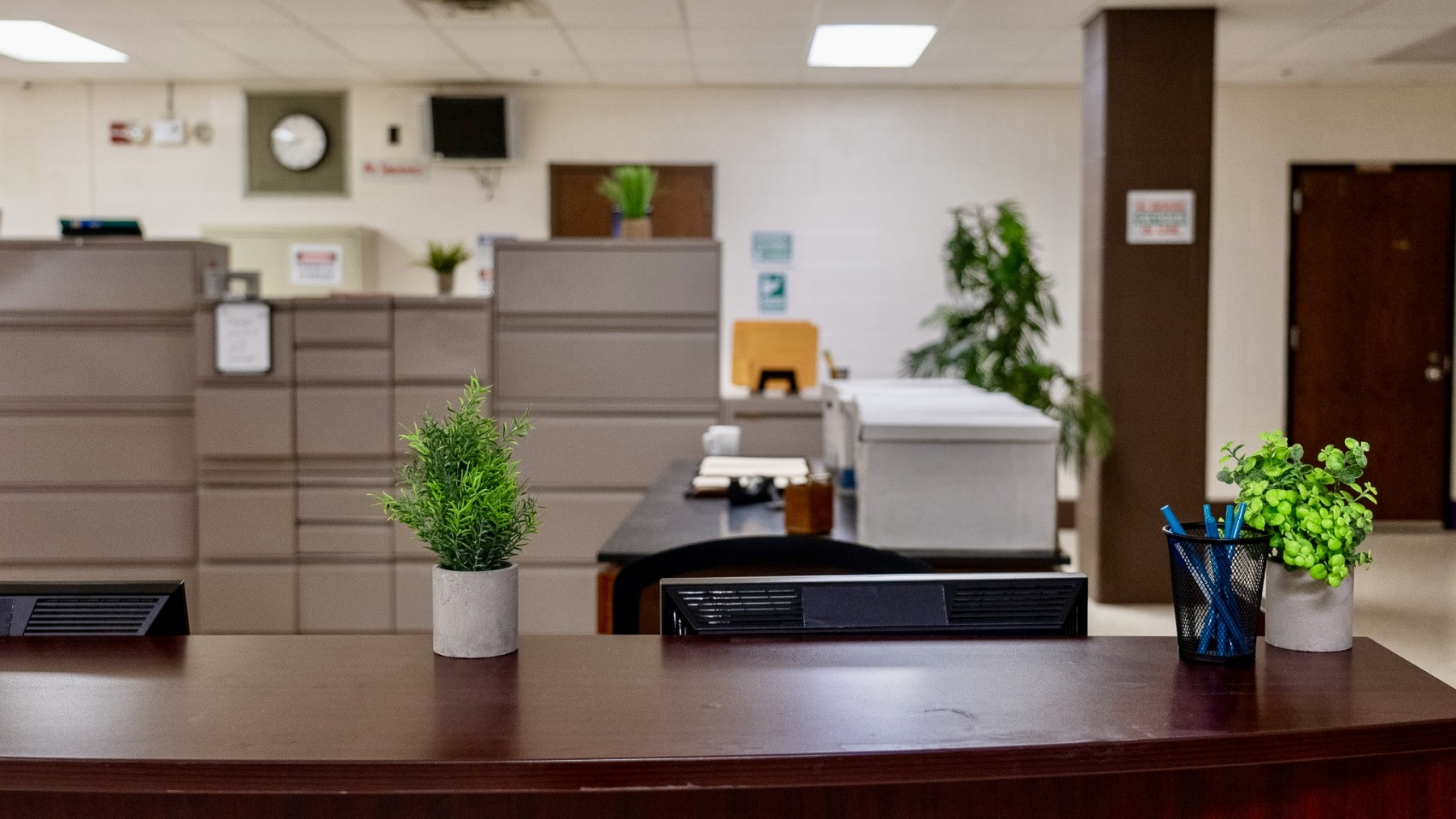 Office reception area with a dark wooden desk featuring small potted plants and a pen holder. Behind the desk are beige divider panels, a printer, and a desk with files. In the background, there are more plants, clock, and signs on a light-colored wa