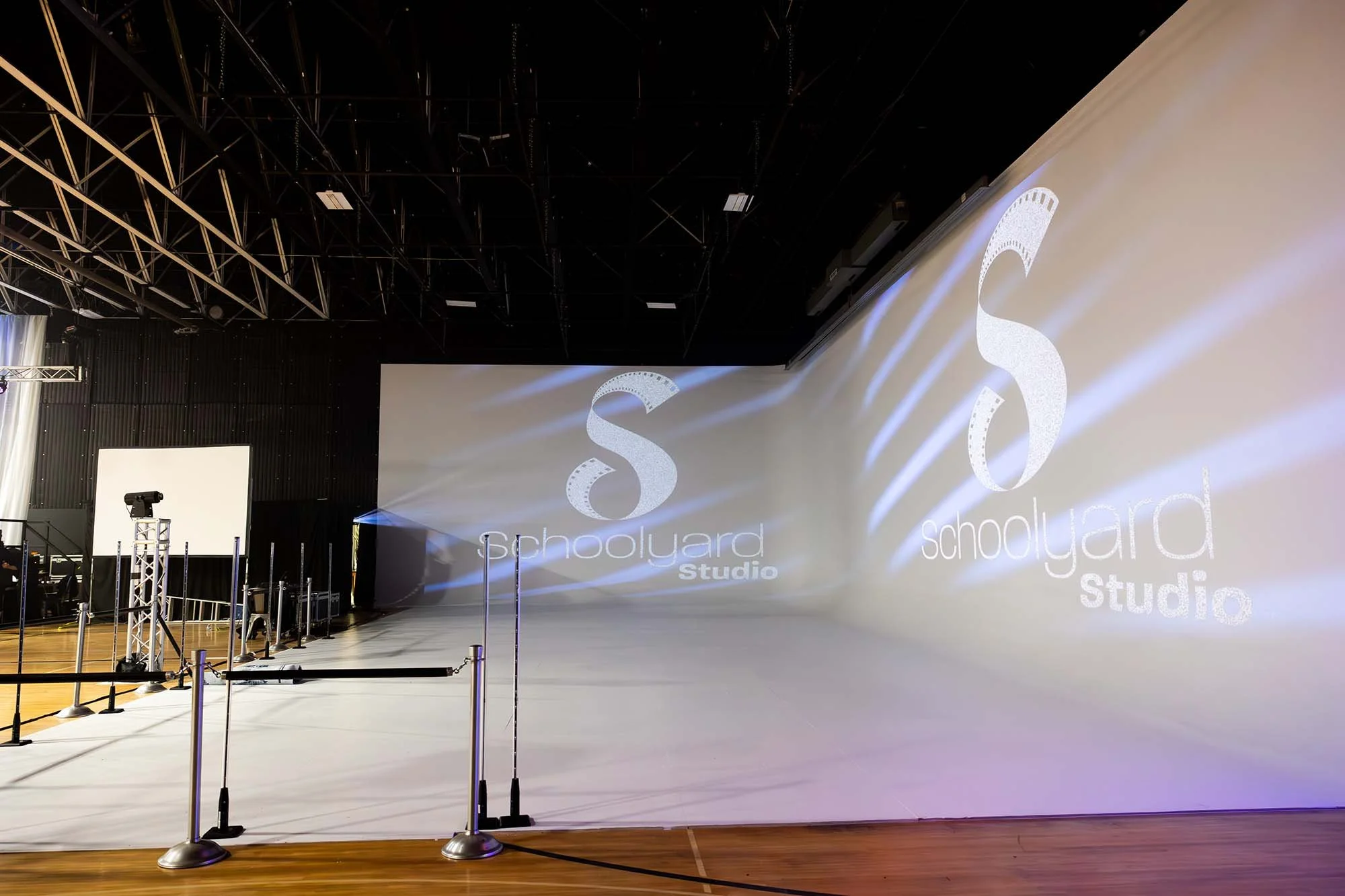 An empty stage with a white backdrop displaying the words "schoolyard studio" and a large letter 'S'. The stage area has barriers and lighting equipment, and the ceiling is black with visible structural elements.