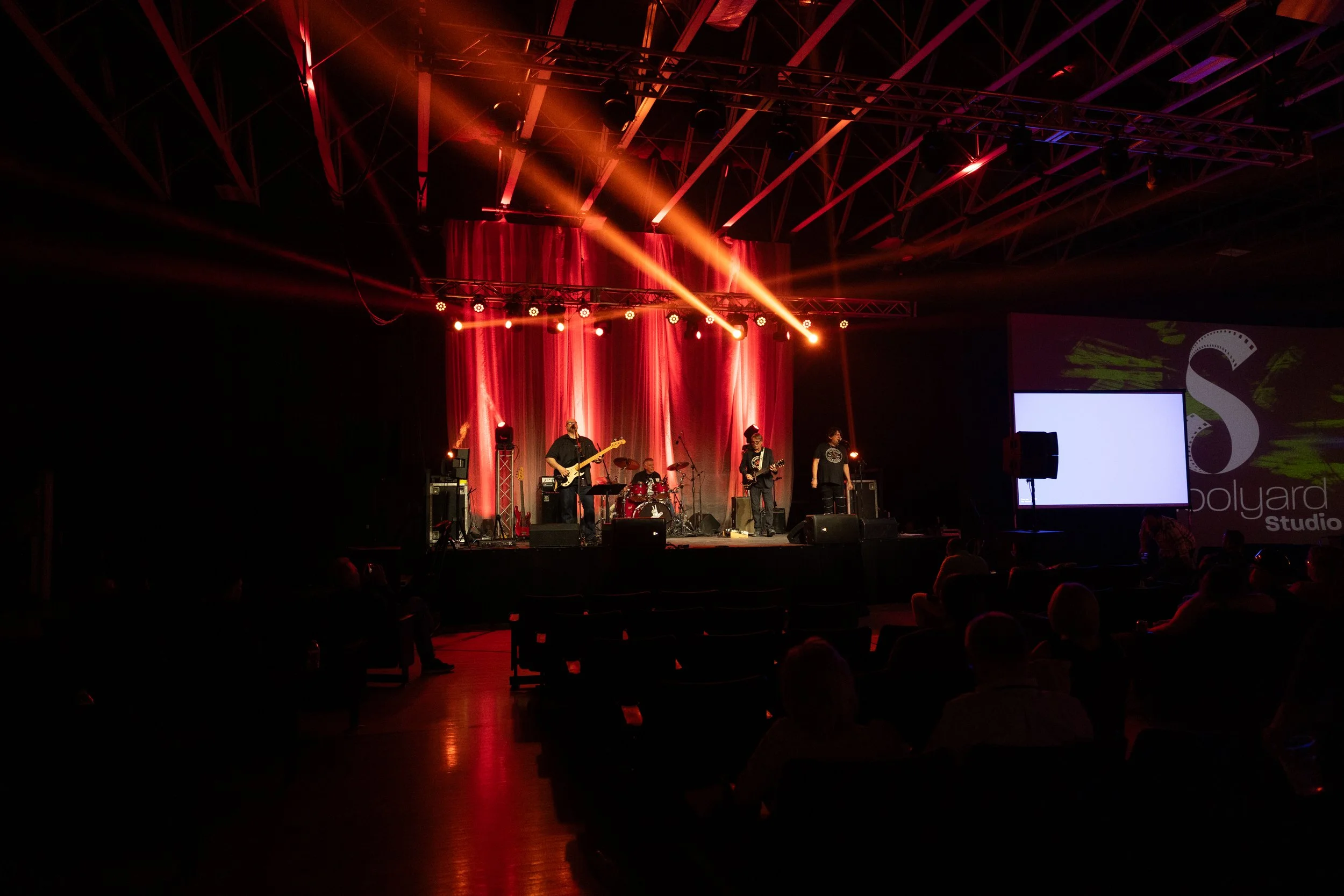A live music performance on a stage with red curtains, four band members playing instruments, and colorful stage lighting in a dark auditorium.
