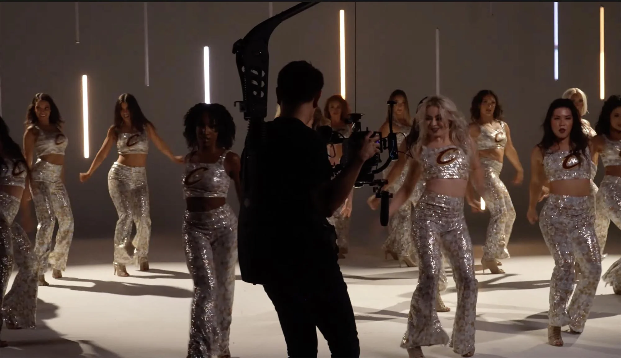 A group of women in shiny, patterned outfits dancing in a studio with vertical light strips on the wall, while a videographer films them.