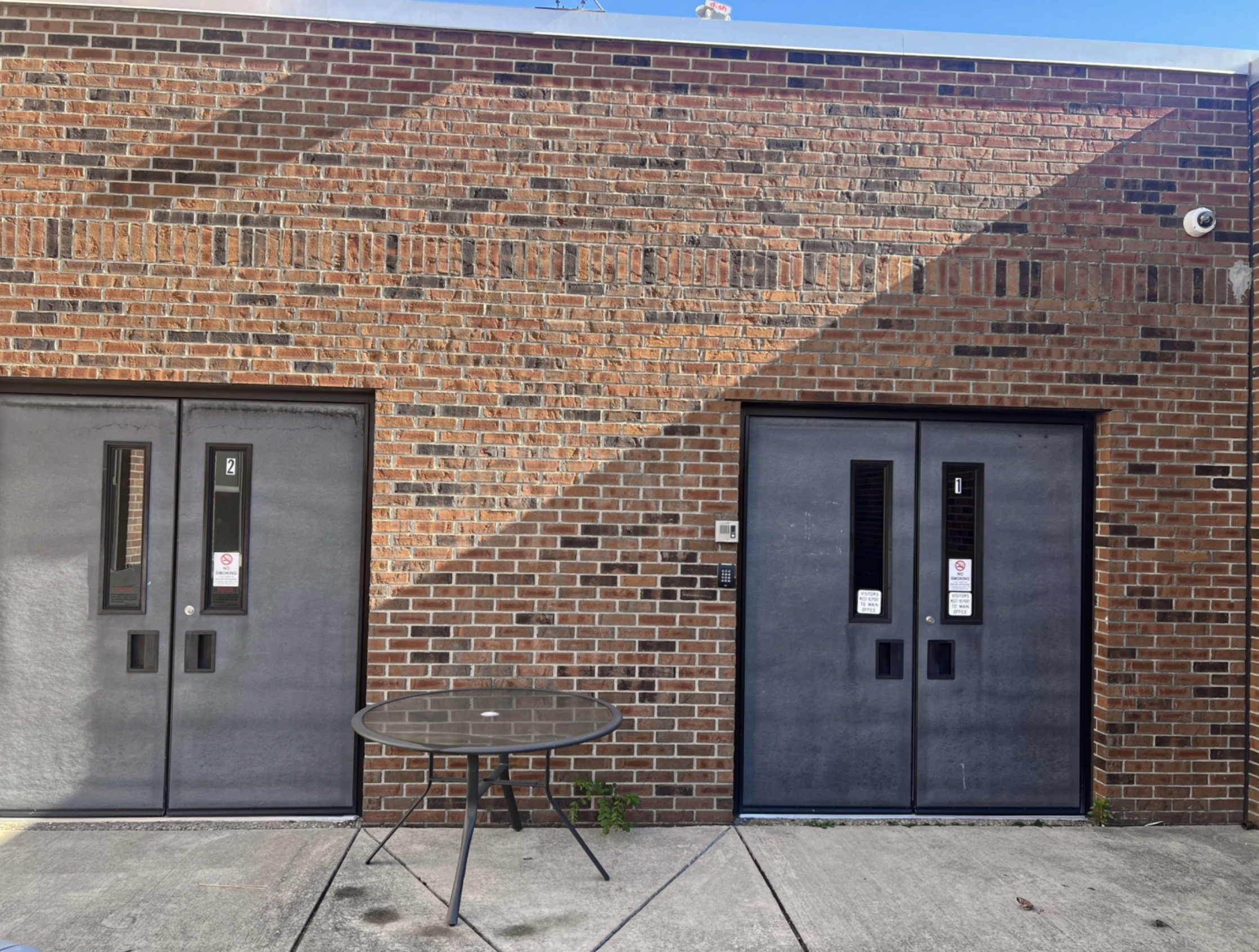 A brick exterior wall with two dark gray metal doors and a small round glass table in front. The wall has a shadow diagonal across it, and there is a security camera in the top right corner.