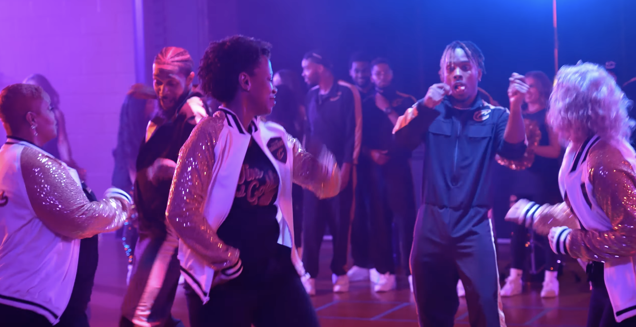 People dancing at a party or nightclub, illuminated by purple and pink lights, with some wearing shiny jackets and casual sportswear.