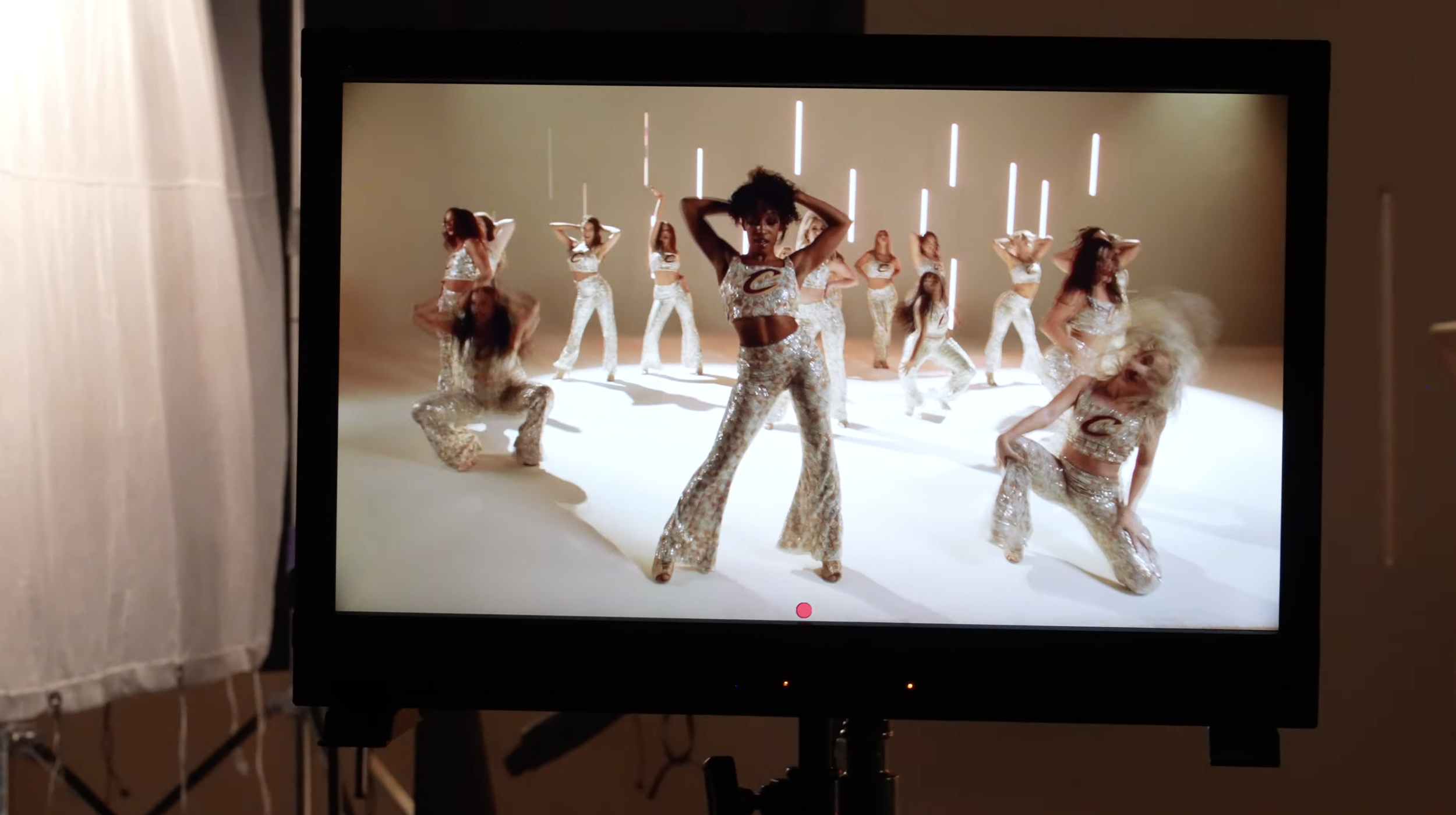 A TV screen displaying a dance performance with multiple women dressed in matching sequined outfits in a studio with neon vertical lights.