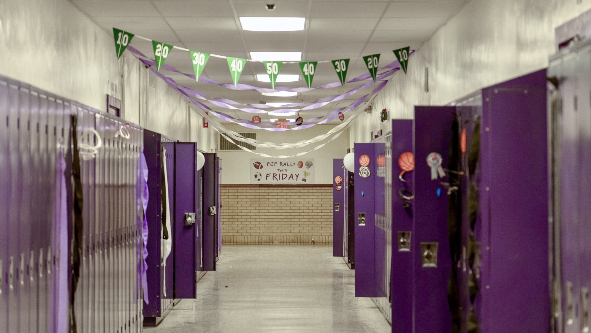A school hallway with purple lockers and purple decorations hanging from the ceiling. A banner at the end of the hallway reads, 'PEP RALLY THIS FRIDAY,' with sports-themed illustrations.
