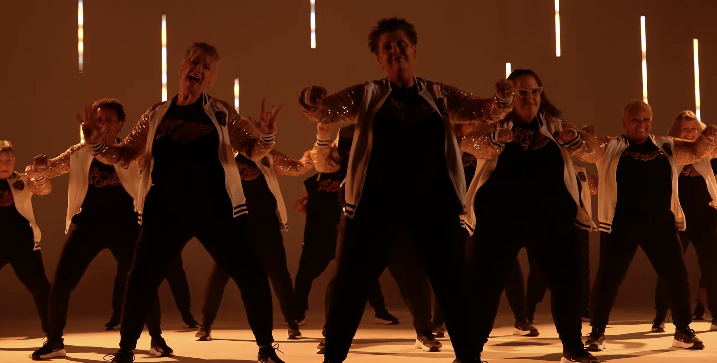 A group of people performing a dance routine on stage, dressed in coordinating outfits with sequined jackets and black pants, with warm stage lighting.