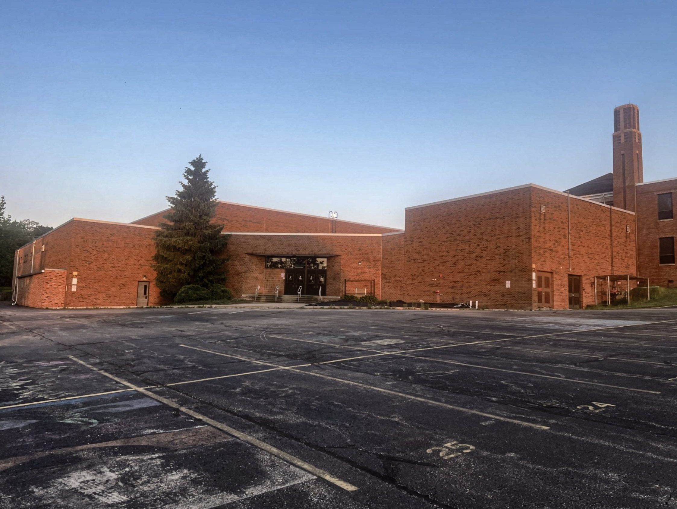 Empty parking lot with cracked asphalt and no cars, in front of a red brick building with a chimney and trees, under a clear blue sky.