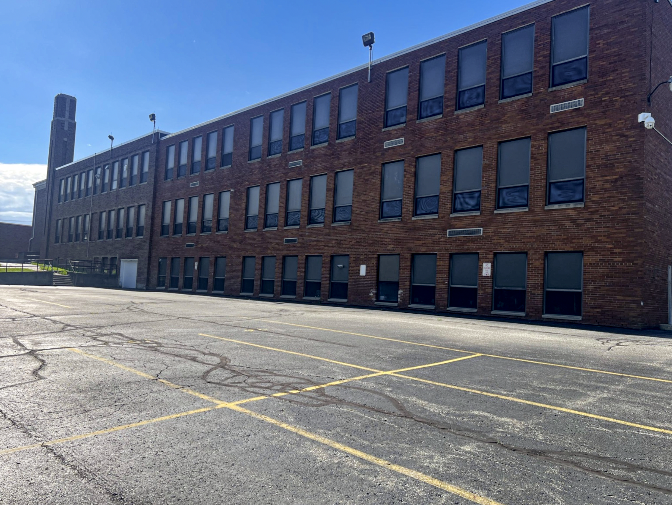 Empty parking lot in front of a brick building with several windows, some covered with shades, and a few security cameras. The sky is clear and blue.