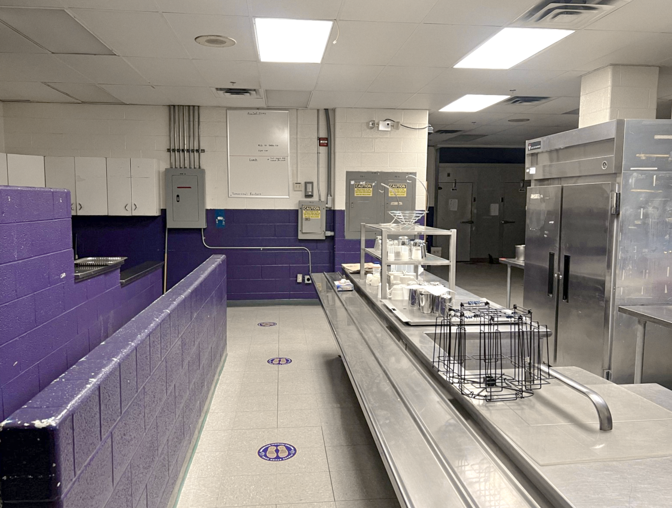 An empty school cafeteria with purple benches, tiled flooring, and kitchen equipment including stainless steel appliances and carts.