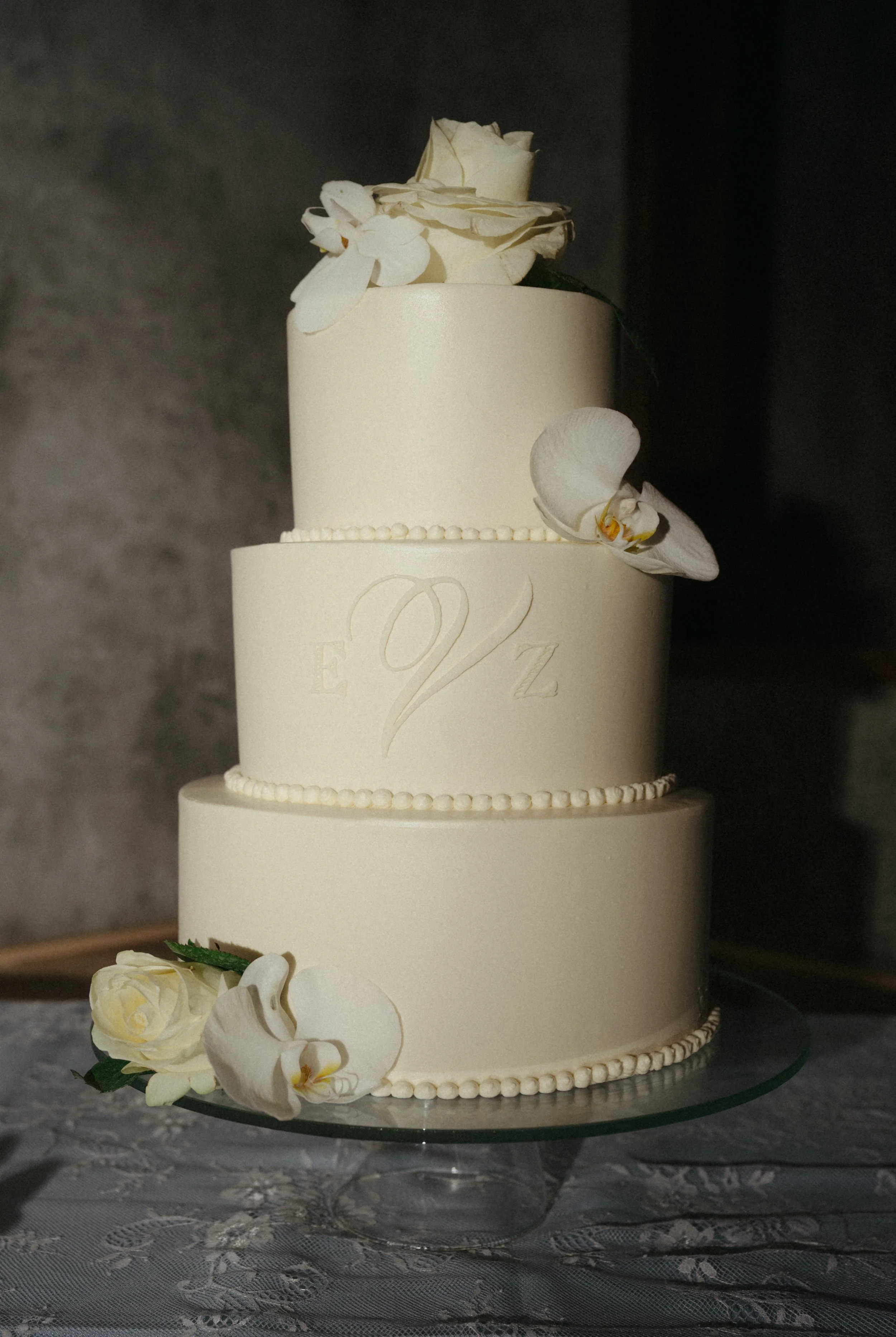 A three-tier white wedding cake decorated with white flowers, sitting on a glass cake stand on a lace tablecloth.