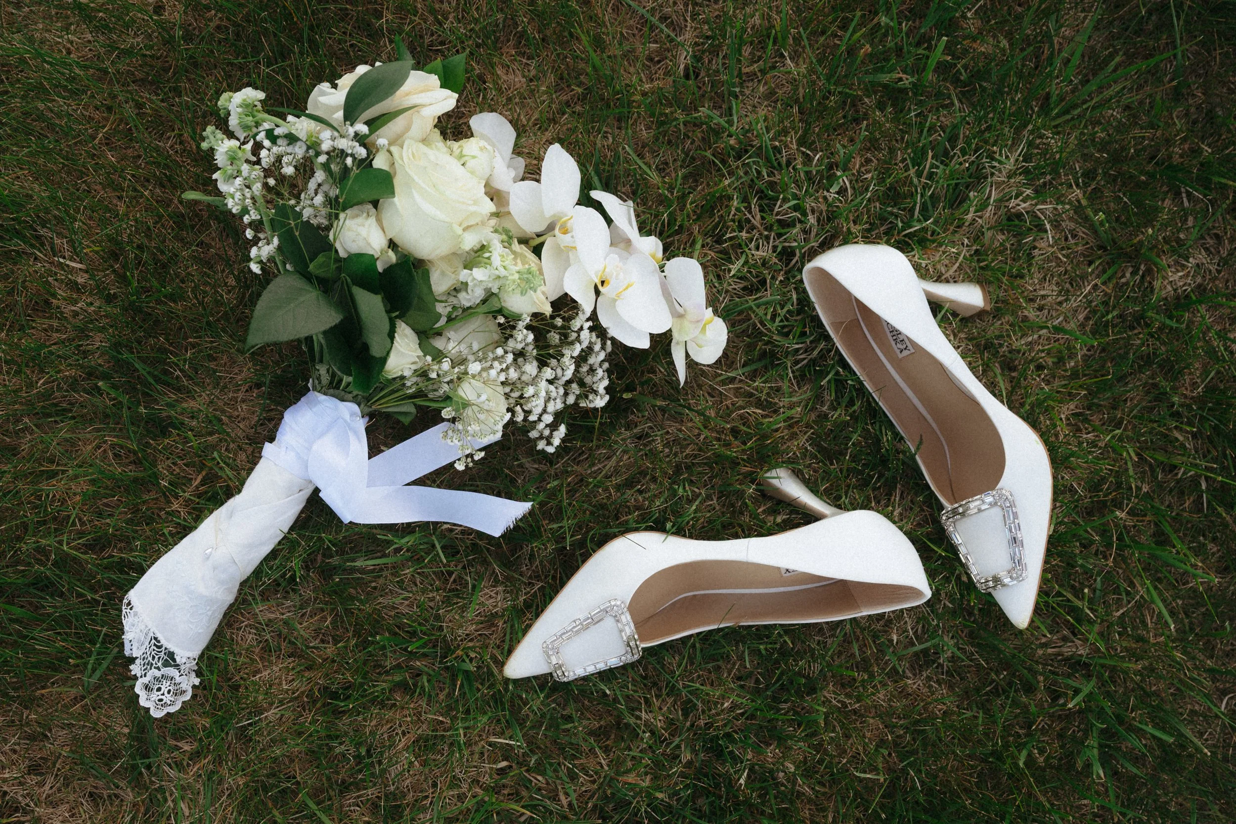 A wedding bouquet of white flowers and greenery, white high-heeled shoes with decorative buckles, and white lace gloves with a ribbon, all lying on grass.