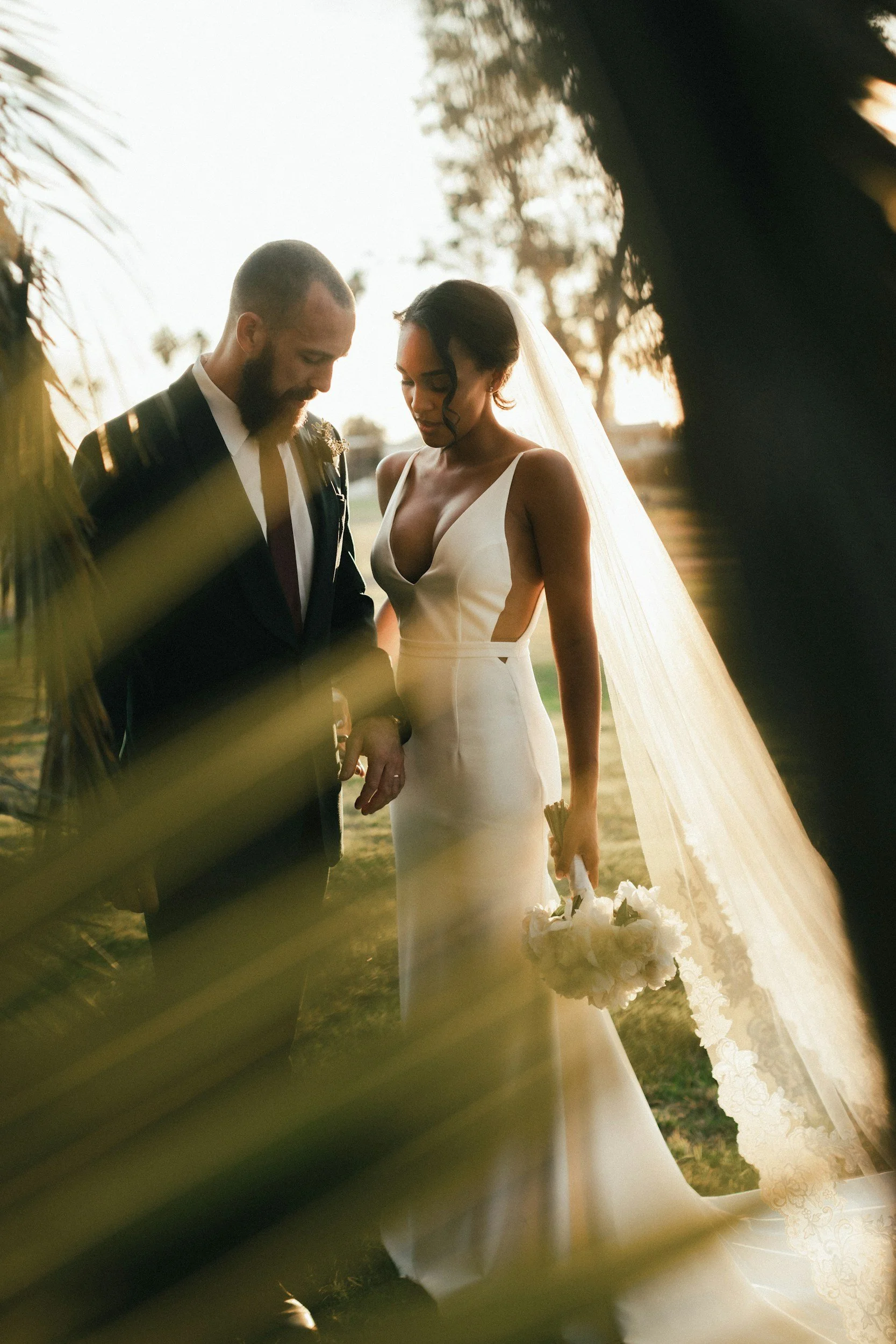 A bride and groom holding hands during their outdoor wedding at sunset, with the bride in a white dress and veil, and the groom in a black suit, framed by tree leaves.