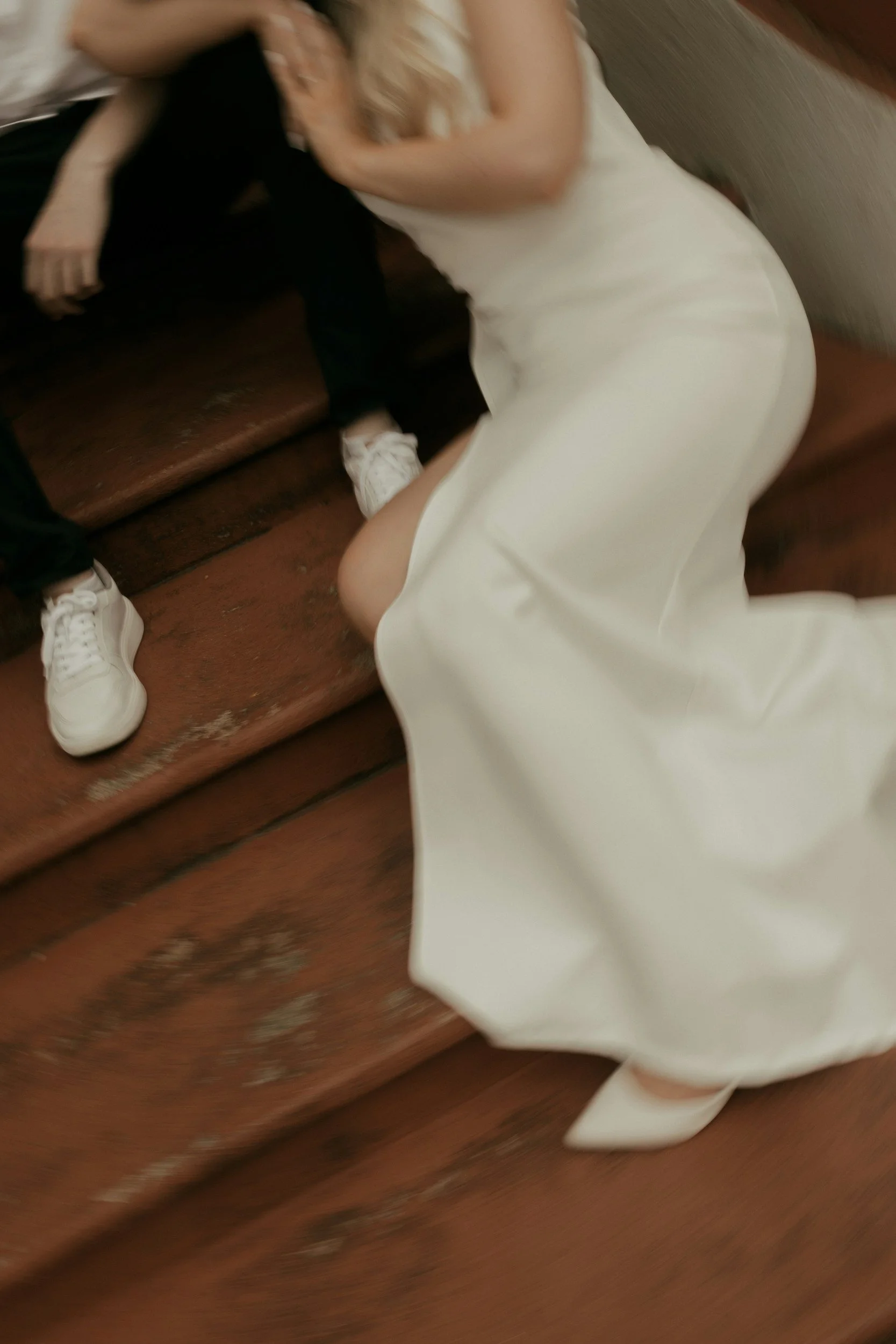 A person in a white dress kneeling on a wooden floor next to a brown leather couch, with another person sitting on the couch wearing black shoes and sportswear.