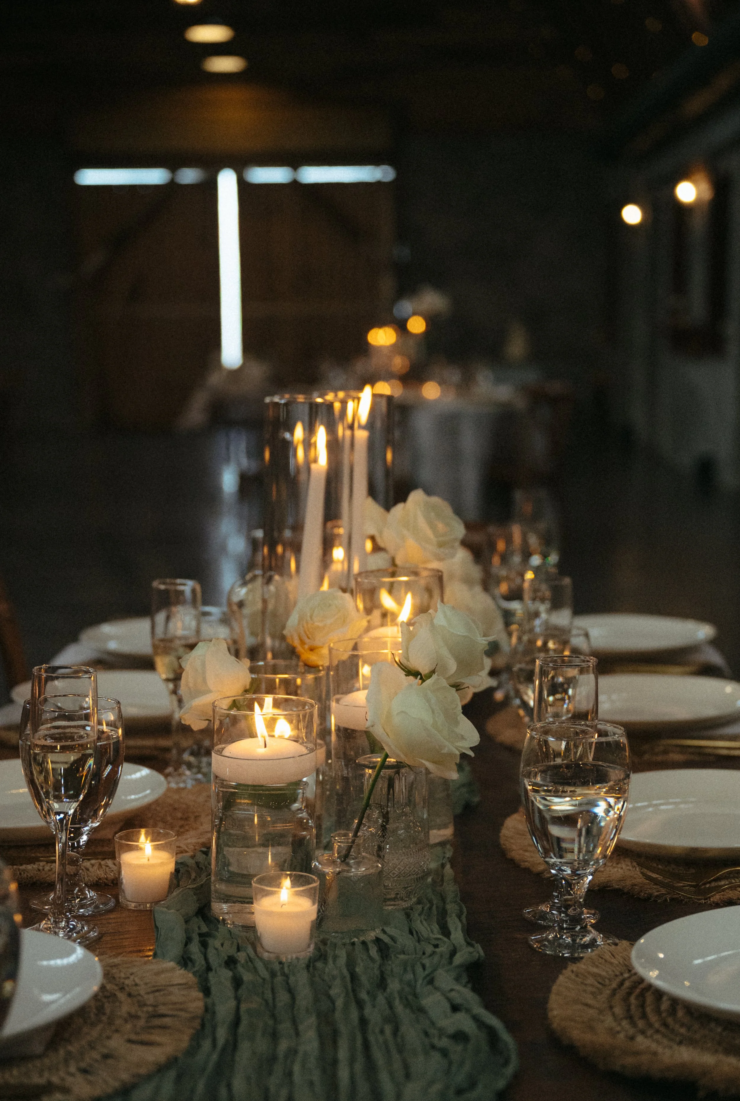 Elegant dining table decorated with white roses and lit candles, set with white plates, glasses, and cutlery, in a dimly lit room.