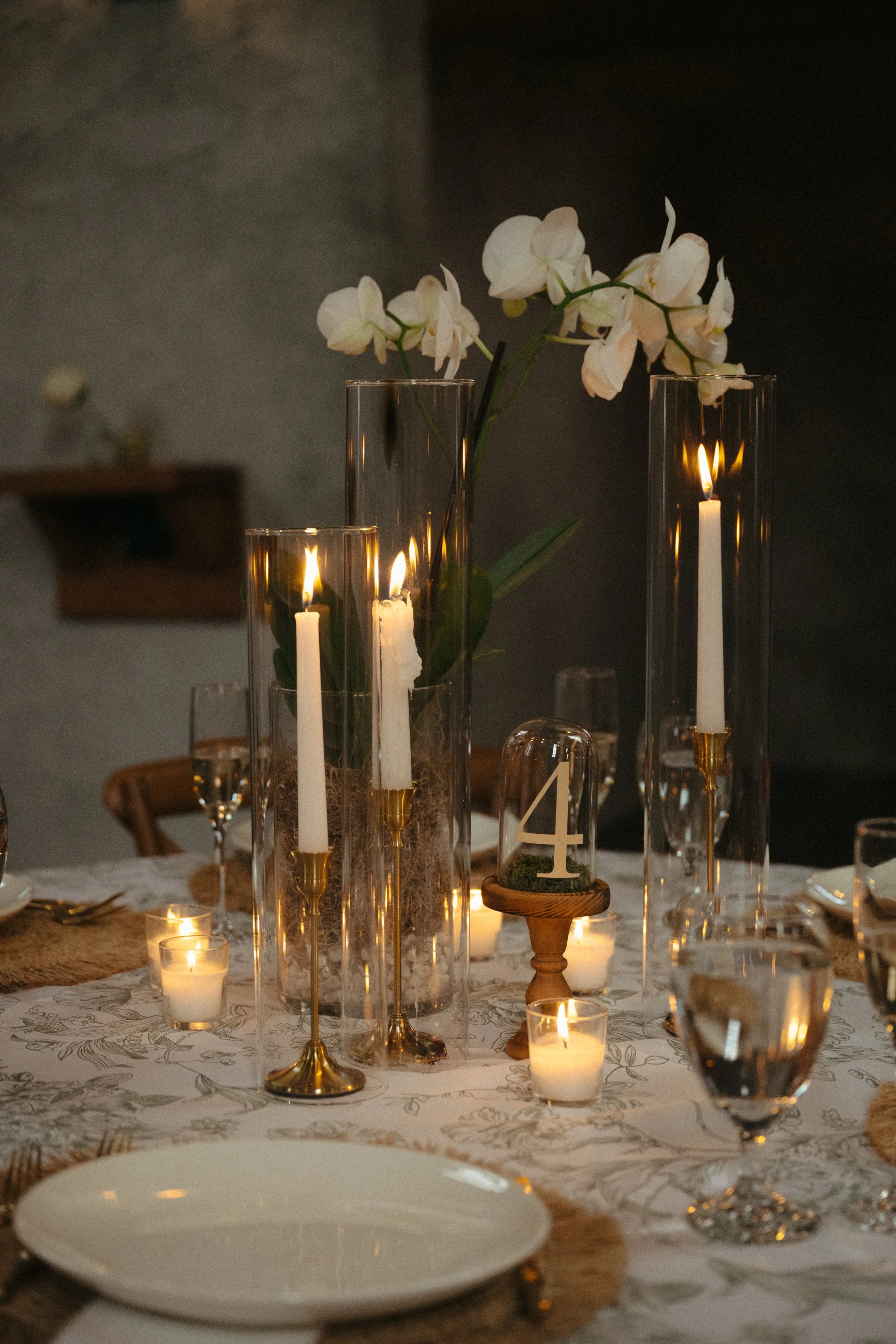 Elegant dining table decorated with tall glass candle holders, white candles, and white orchids, with warm candlelight creating a cozy atmosphere.