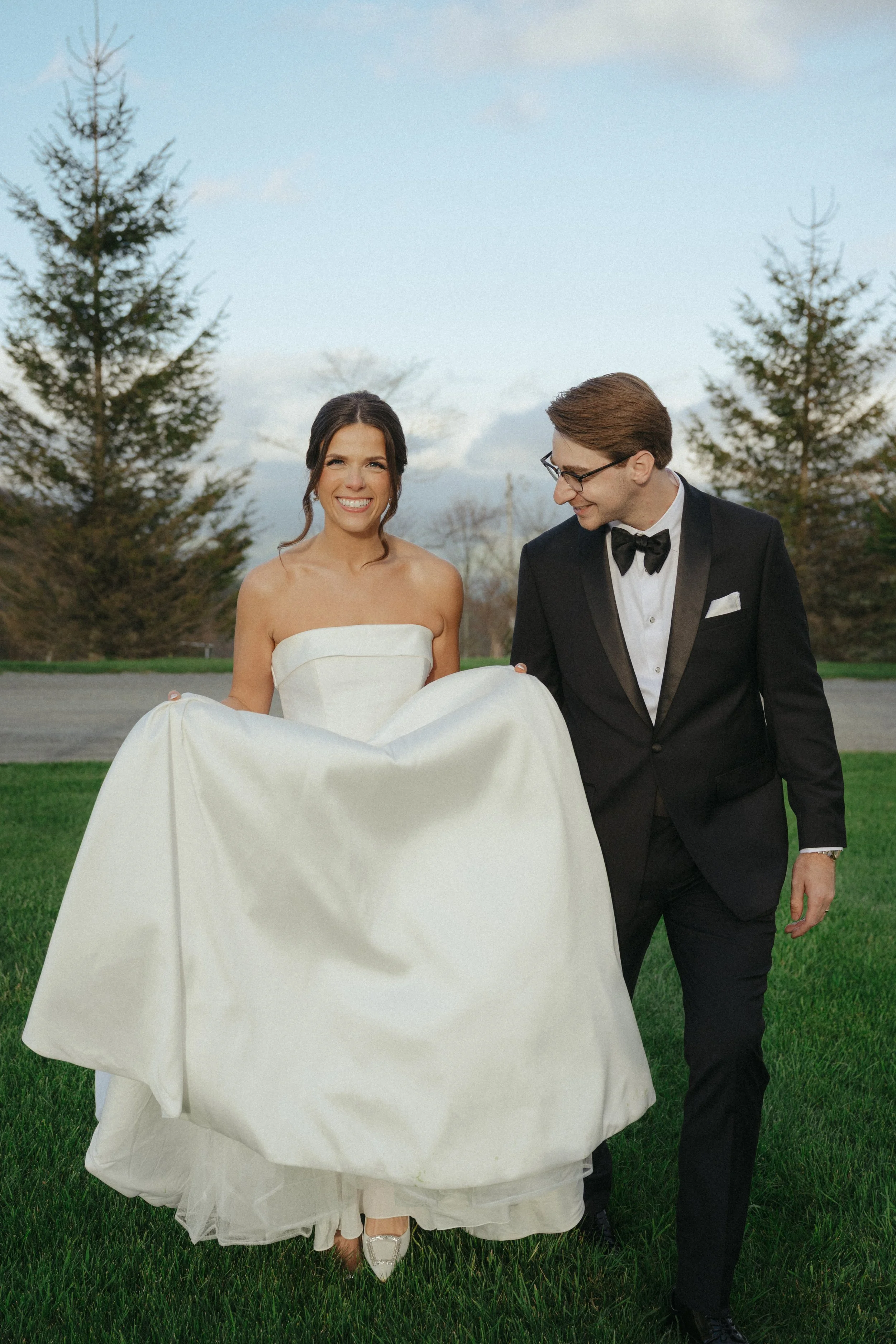 A bride and groom walking outdoors on grass, the bride in a strapless wedding dress holding her dress with both hands, and the groom in a black tuxedo with a bow tie, smiling and looking at each other, with trees and a cloudy sky in the background.