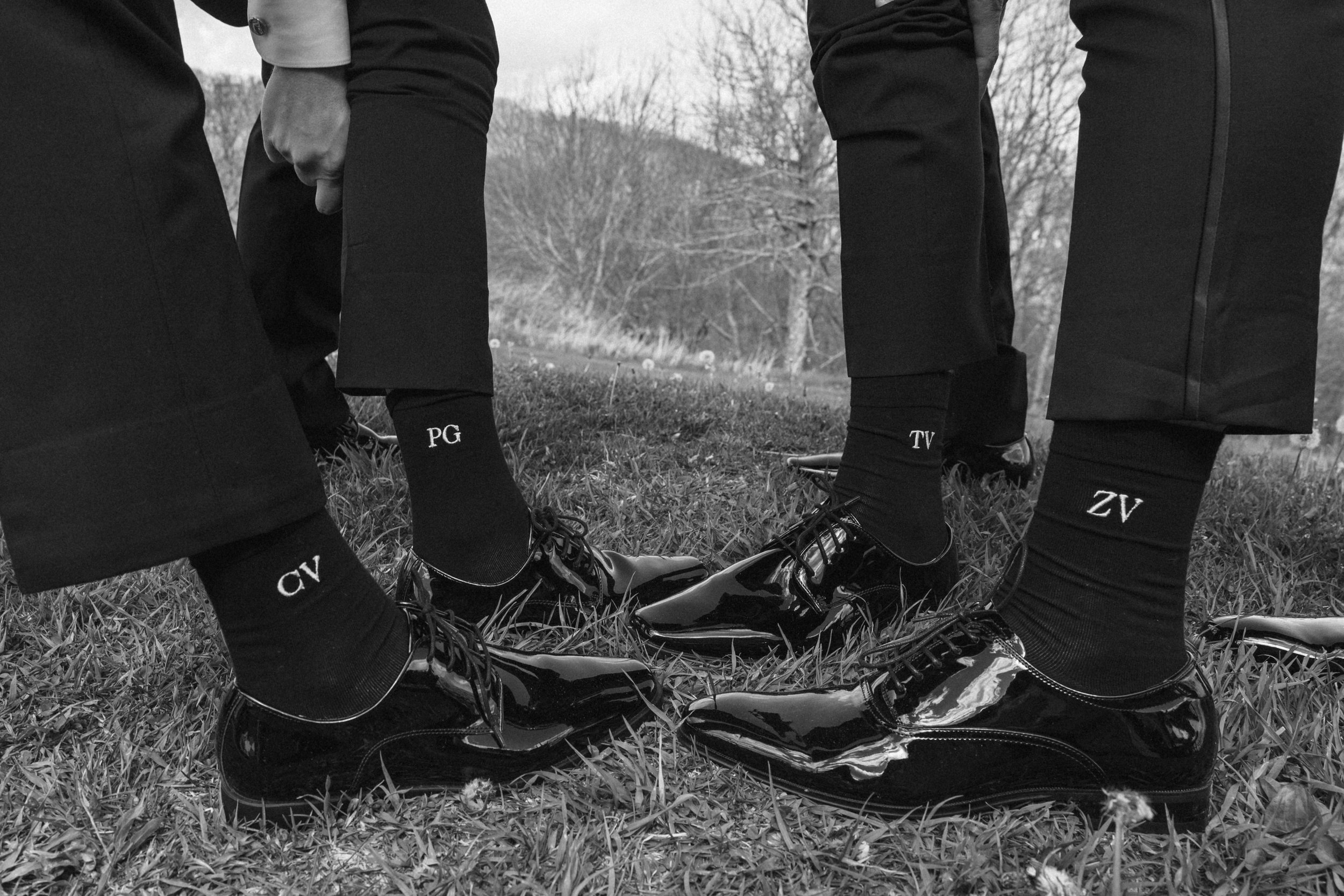 Close-up of three people wearing formal business suits and shiny black dress shoes, standing on a grassy field outdoors, with their legs slightly apart and hands in pockets.