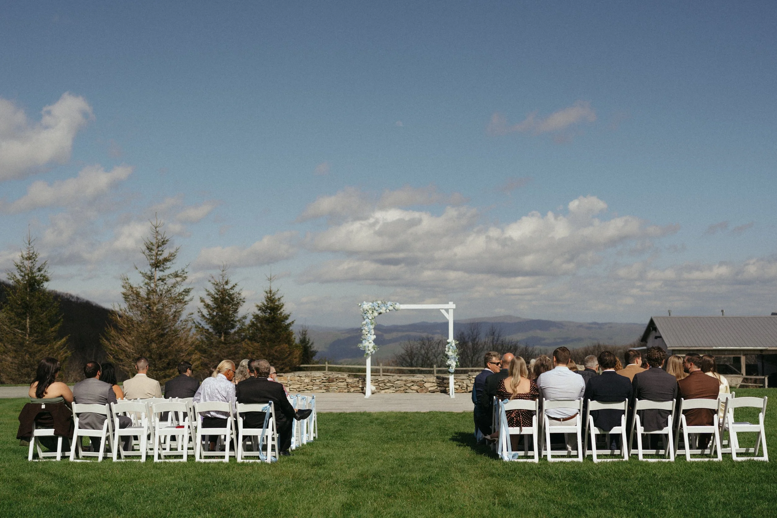 Outdoor wedding ceremony with guests sitting in white chairs, decorated arch, and scenic mountainous background under a partly cloudy sky.