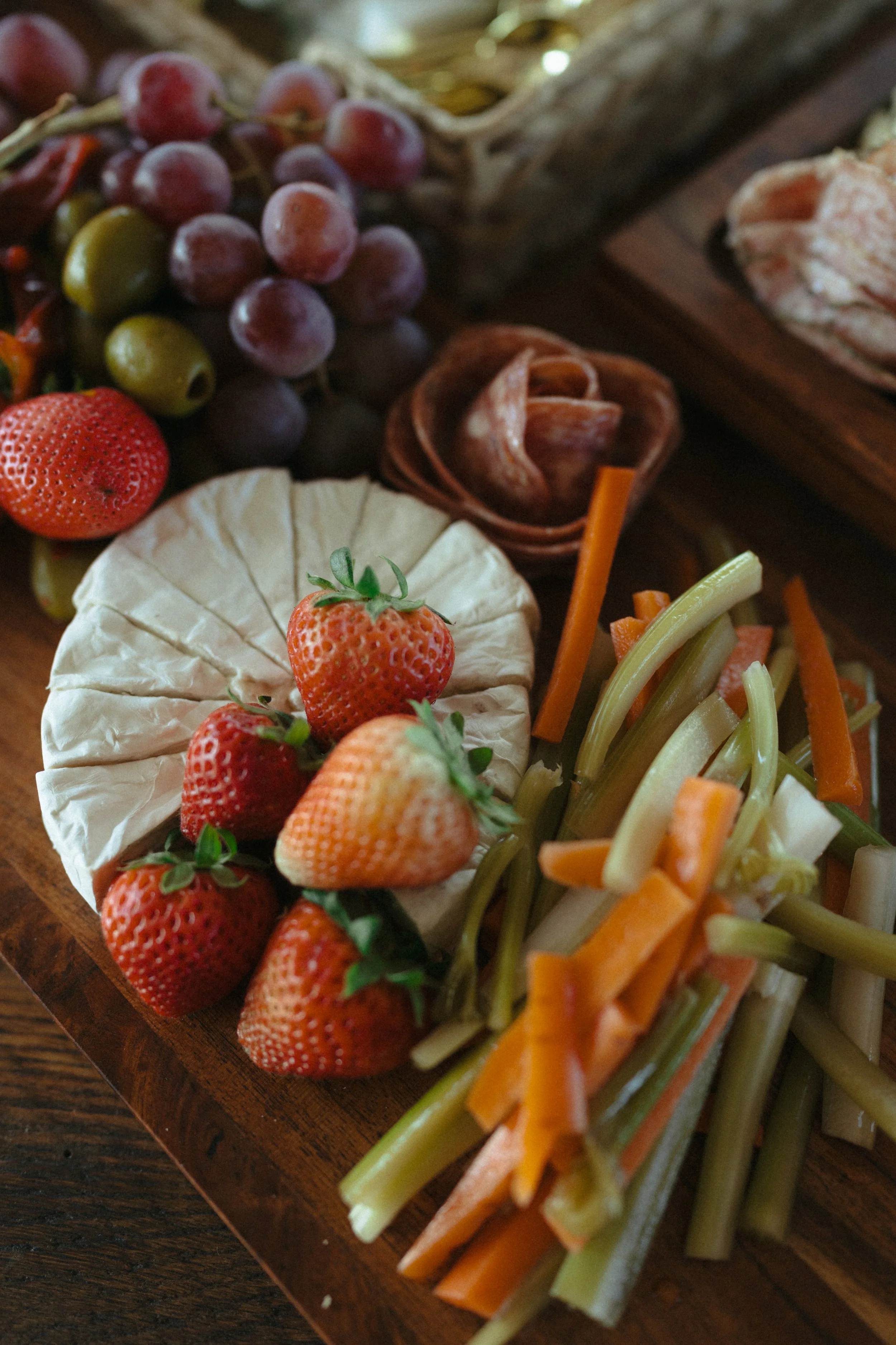 A wooden platter with fresh strawberries, grapes, cheese, sliced vegetables, and cured meat.