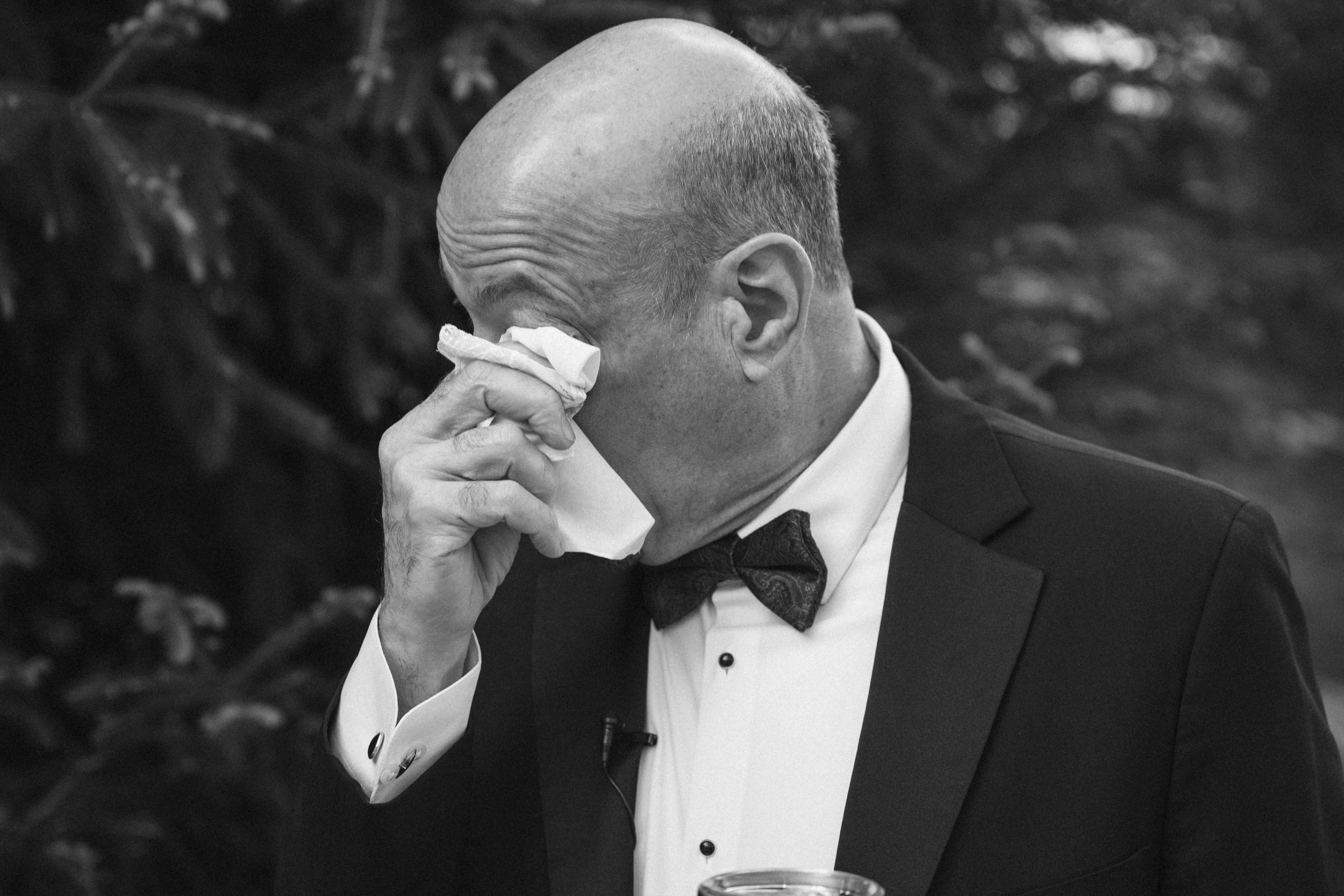 A black and white photo of a man dressed formally in a tuxedo, adjusting his earpiece with a napkin. He has a bald head and is wearing a bow tie. The background is blurred with some foliage.