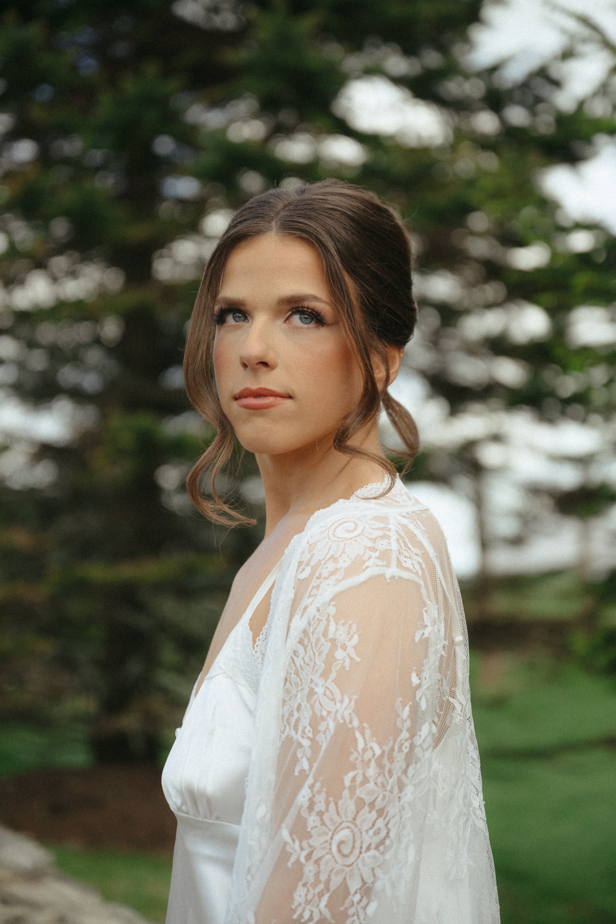 A woman with brown hair styled in loose waves, wearing a white lace dress with intricate patterns, standing outdoors in front of a blurred background of trees with green foliage.