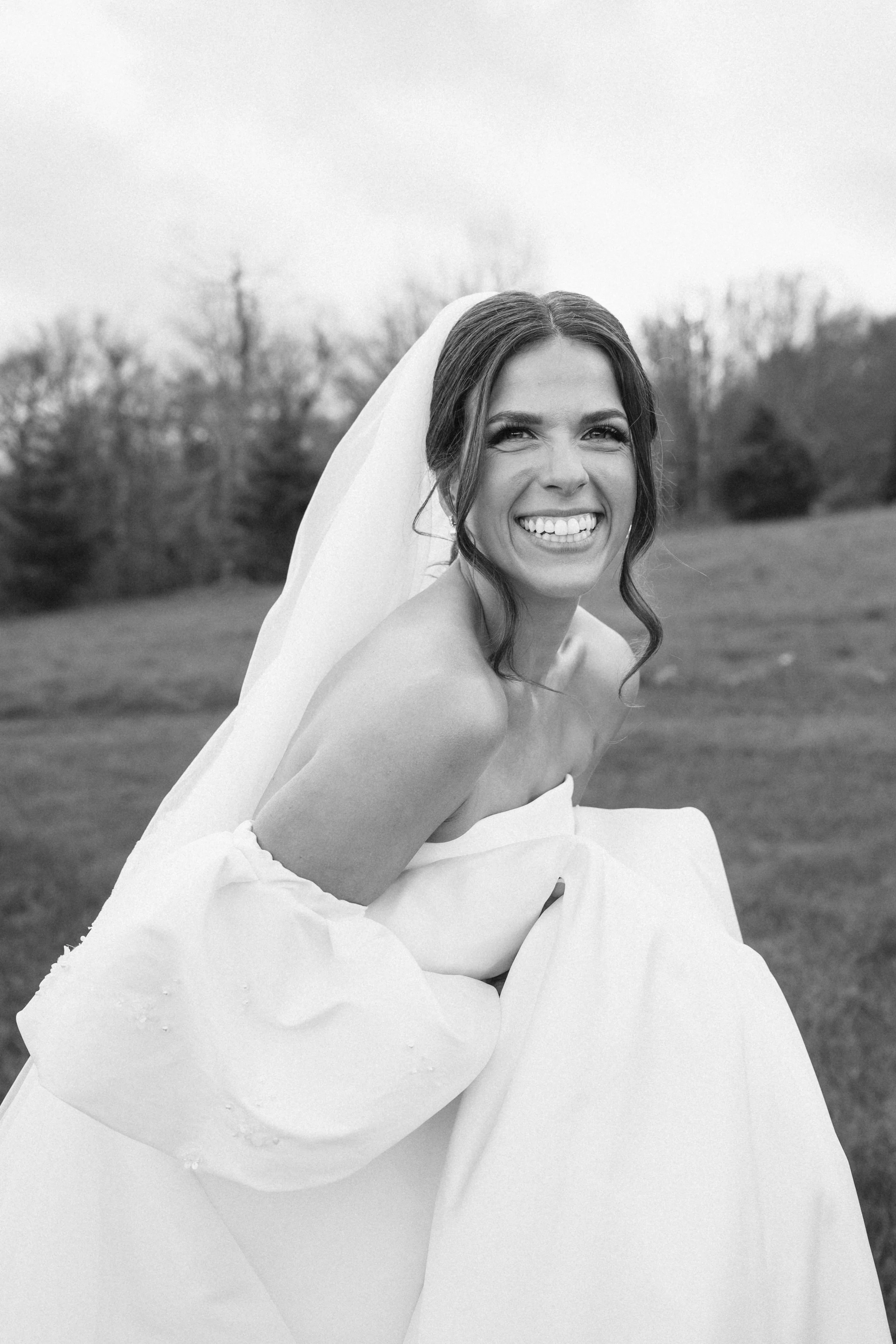 A bride with a smile, wearing a strapless wedding dress and veil, outdoors with trees in the background.