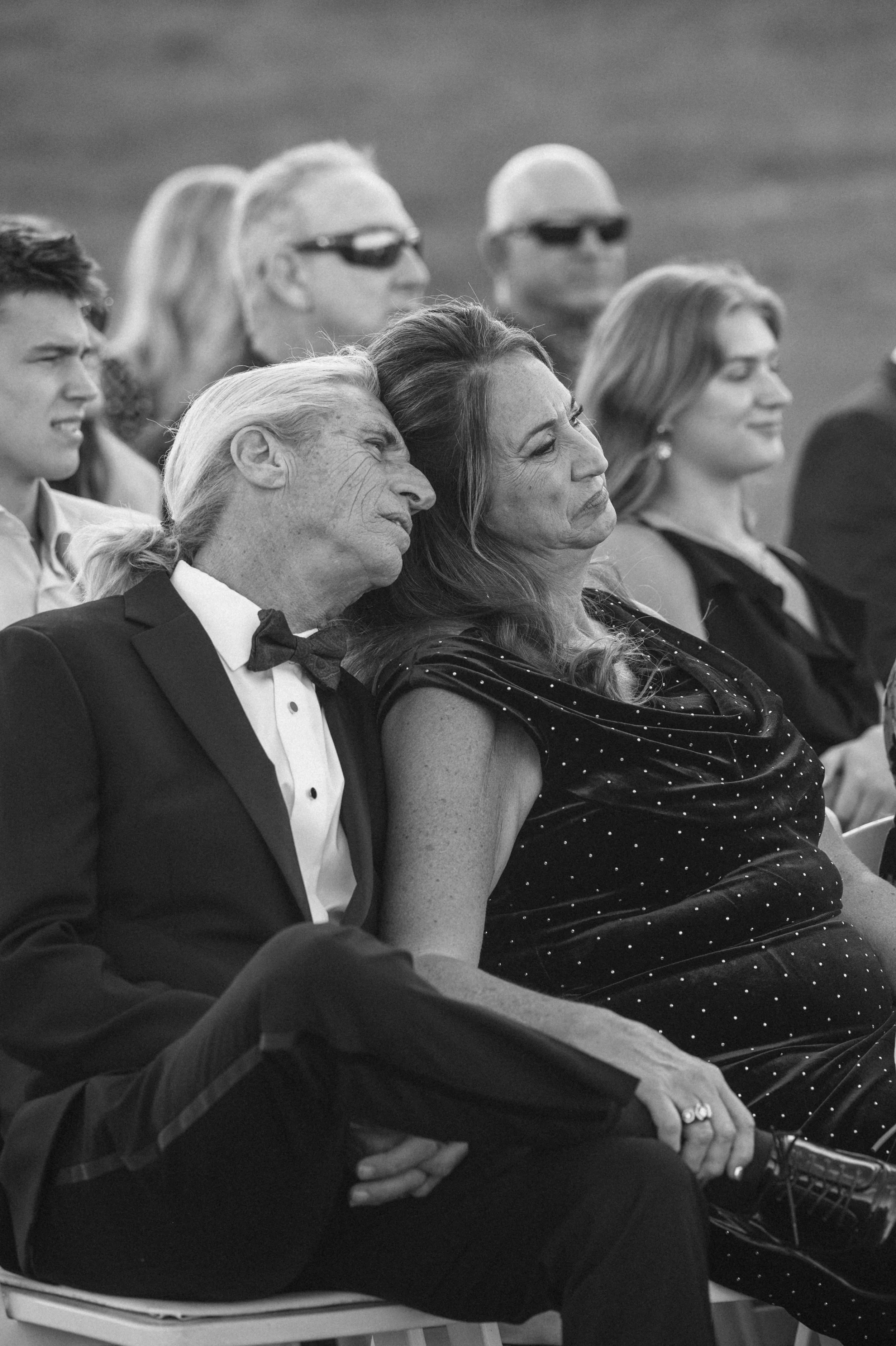 Black and white photo of people seated in an audience, including older man and woman in the front row, with the man resting his head on the woman's shoulder, and background showing others wearing sunglasses and formal attire.