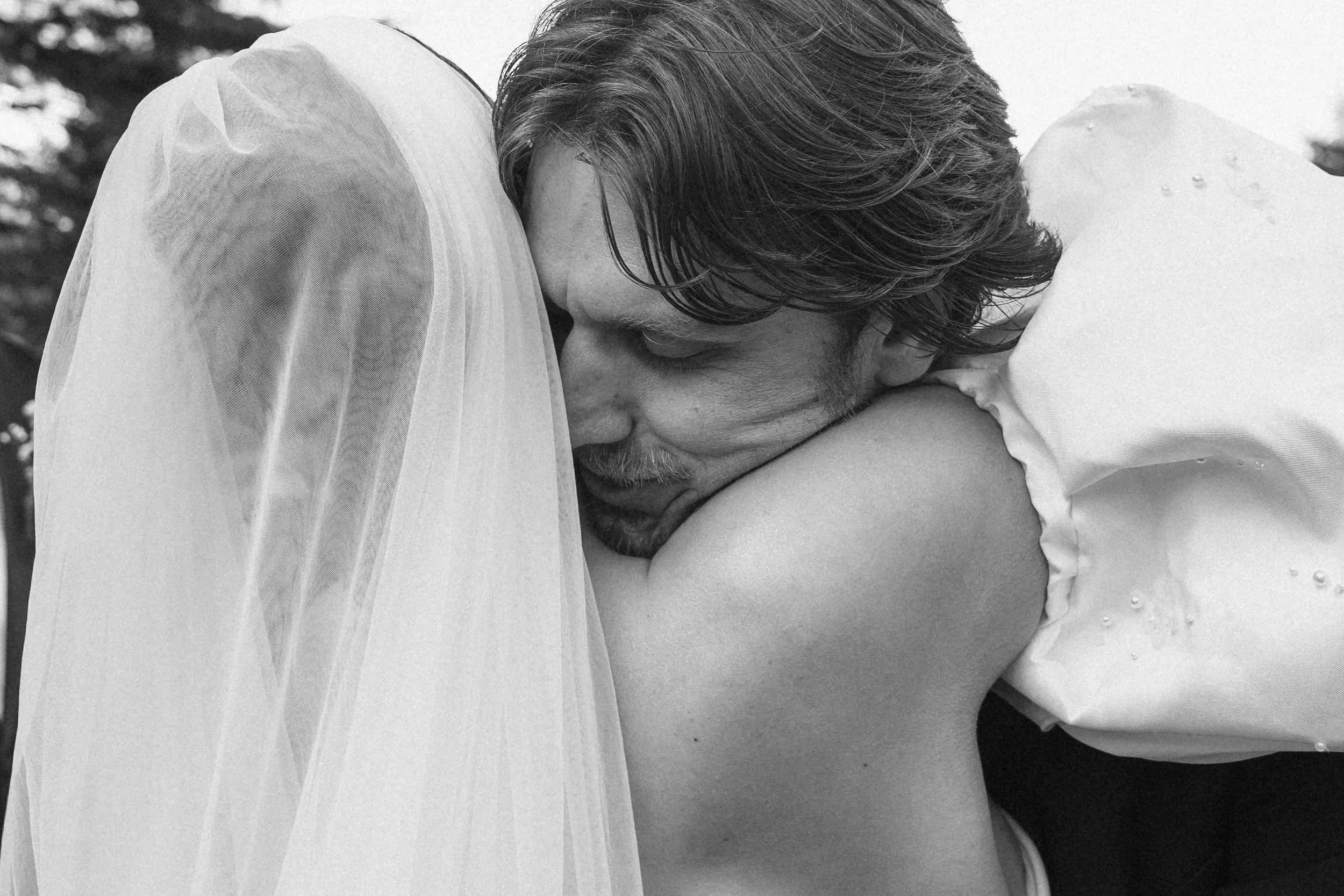 A black and white photograph of a man with medium-length hair and a mustache, embracing and resting his head on a woman's shoulder, who is wearing a white garment with a sheer overlay.