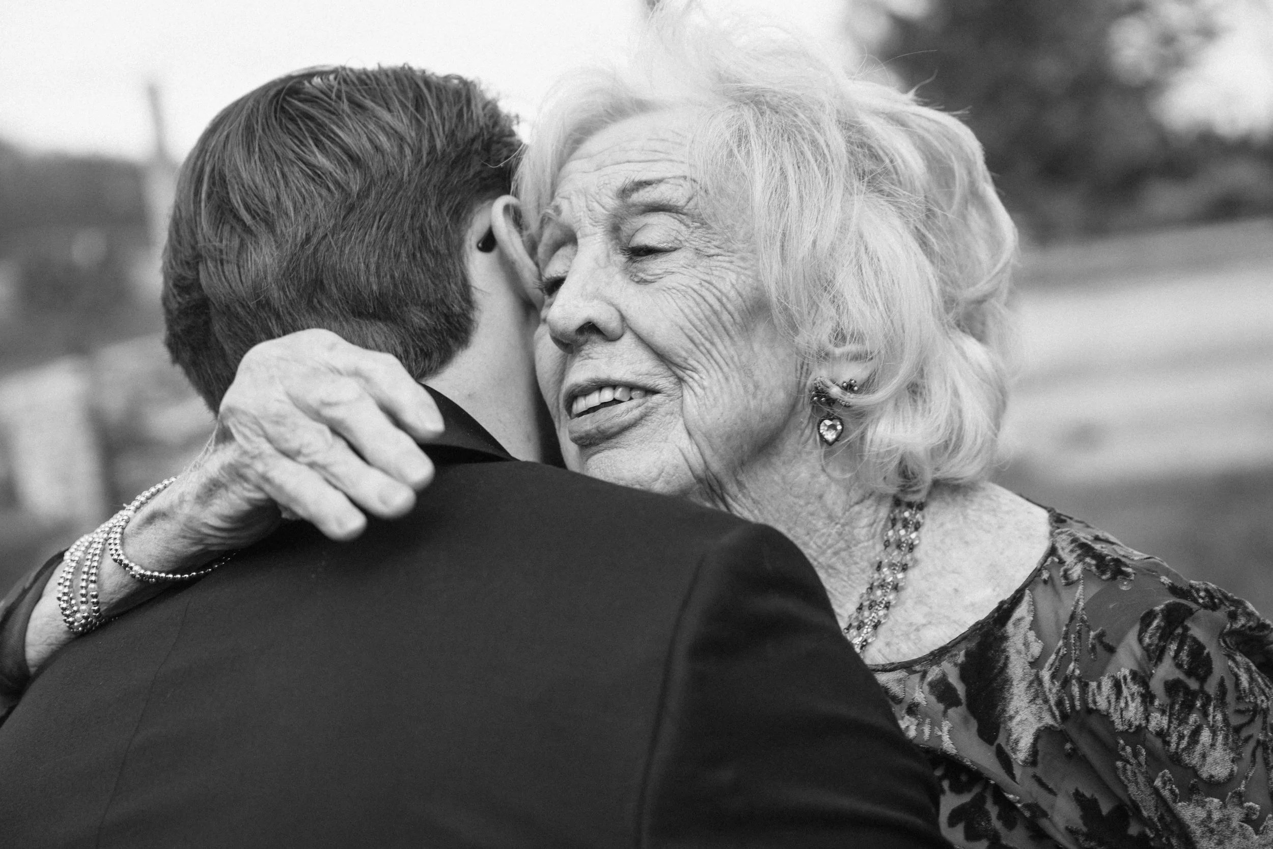 An elderly woman with curly hair and earrings embracing a man in a suit, closing her eyes with a content expression on her face.