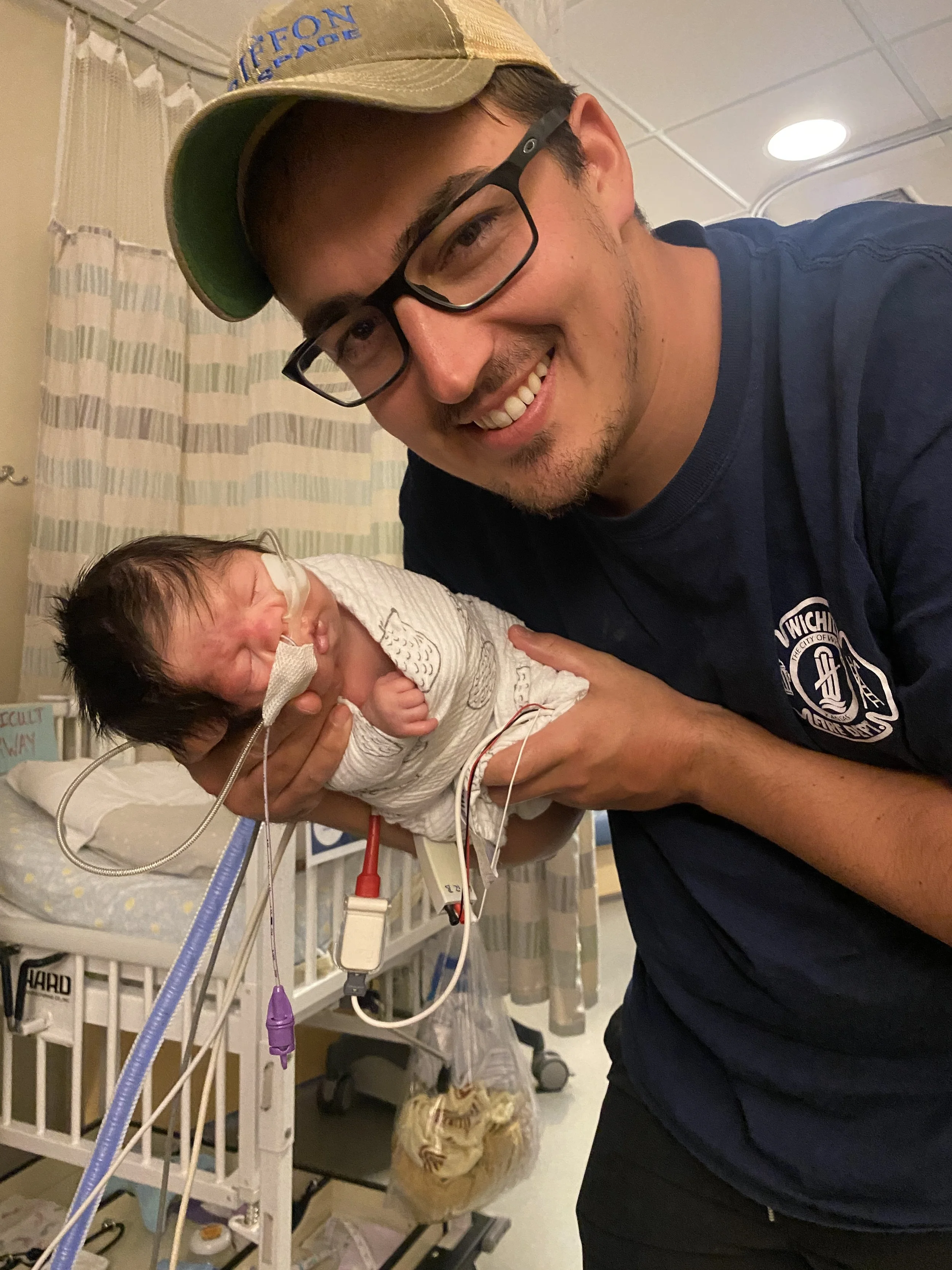 A man with glasses and a beige cap smiling while holding a newborn baby in a hospital room. The newborn has a nasal cannula, is wrapped in a blanket, and has wires attached.