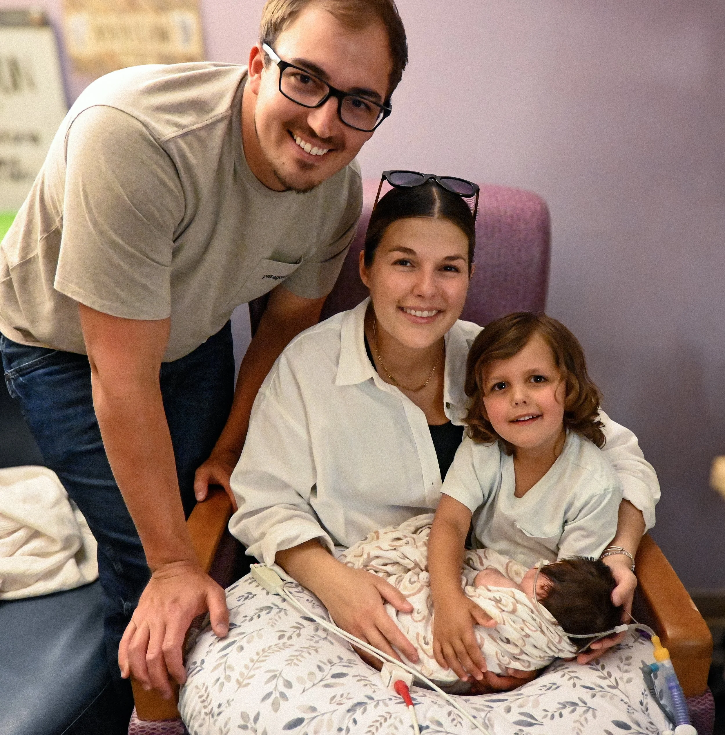 A young girl with brown hair lying in a hospital bed, connected to medical tubes, with a woman, and a man with glasses and short brown hair, smiling and leaning over her.