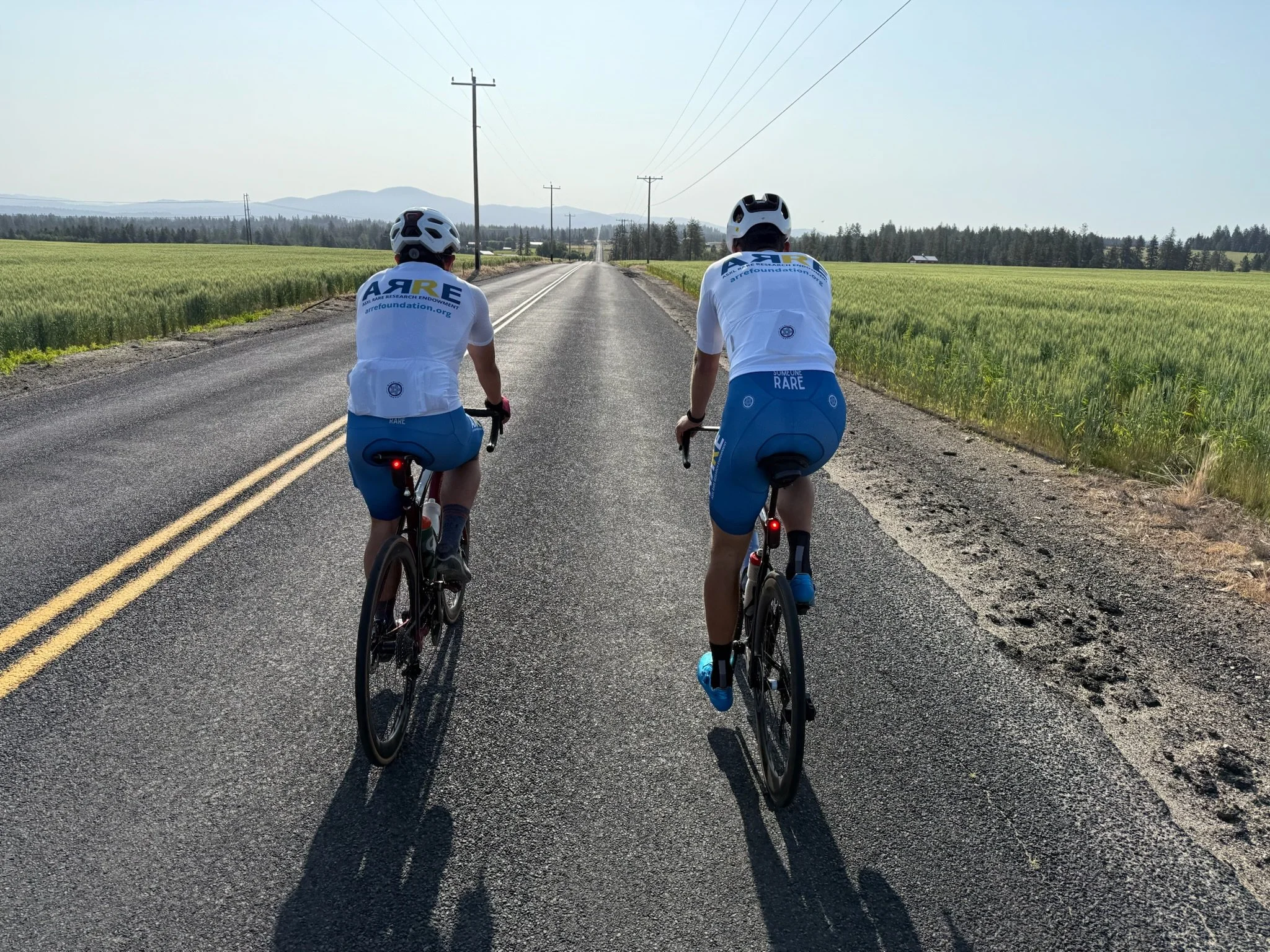Two cyclists wearing white helmets and matching white cycling jerseys ride down a rural road lined with fields of green crops, with telephone poles extending into the distance on a sunny day.