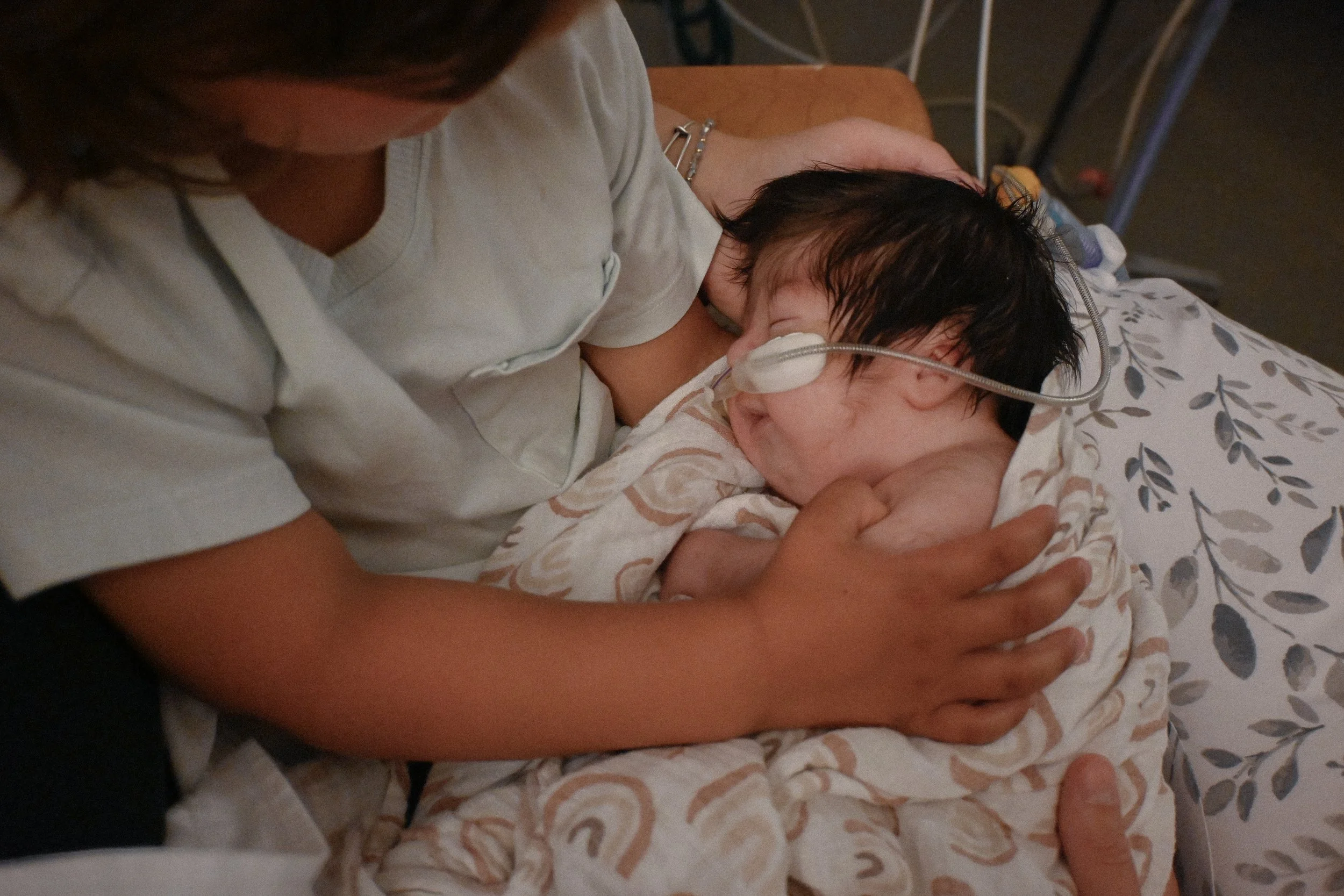 A woman holding a crying newborn baby with a nasal cannula for oxygen, sitting on a bed with a leaf-patterned blanket.