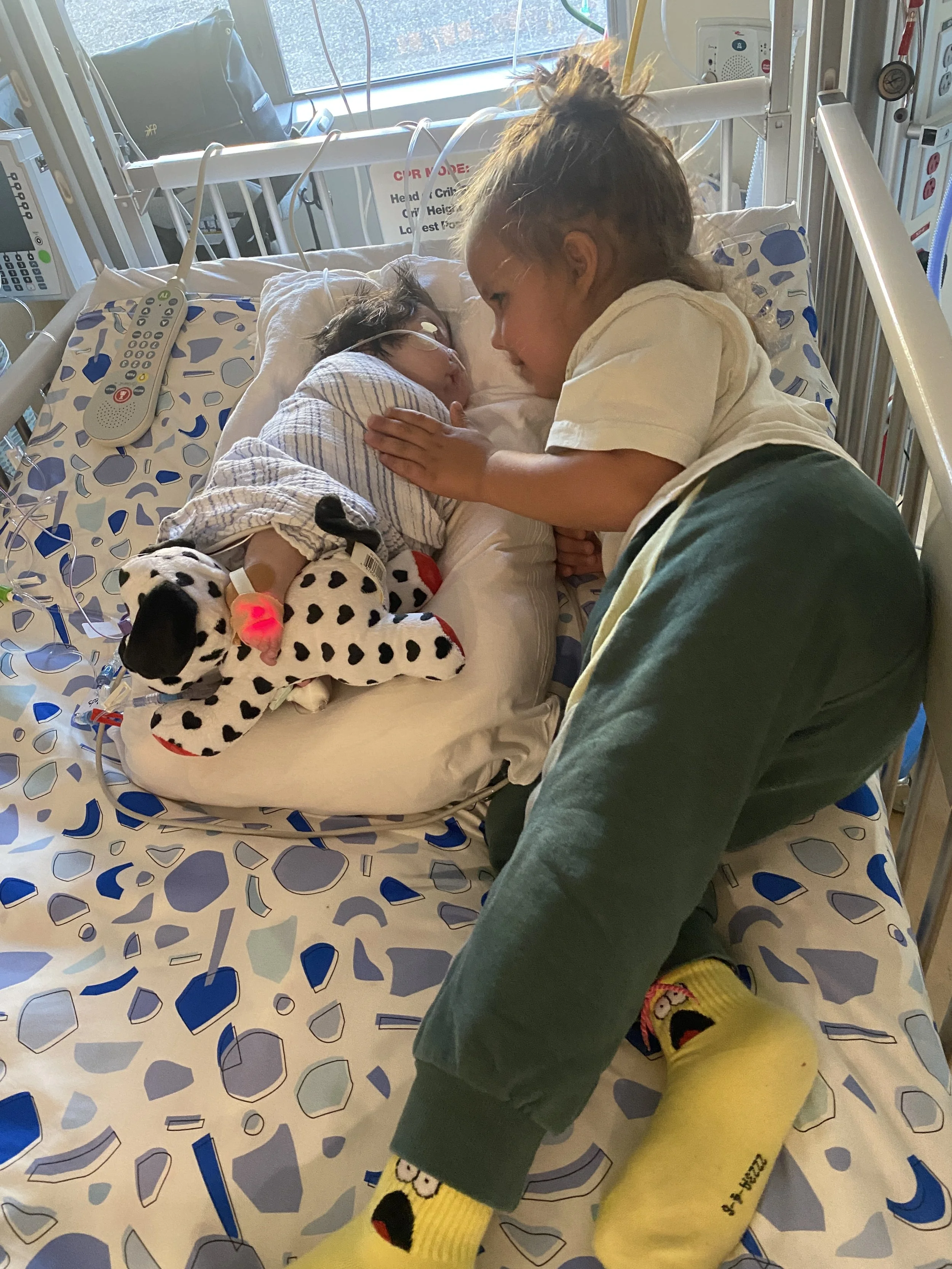 A young girl in hospital bed with a feeding tube, being gently touched and comforted by an older child, in a hospital room with medical equipment and a window in the background.