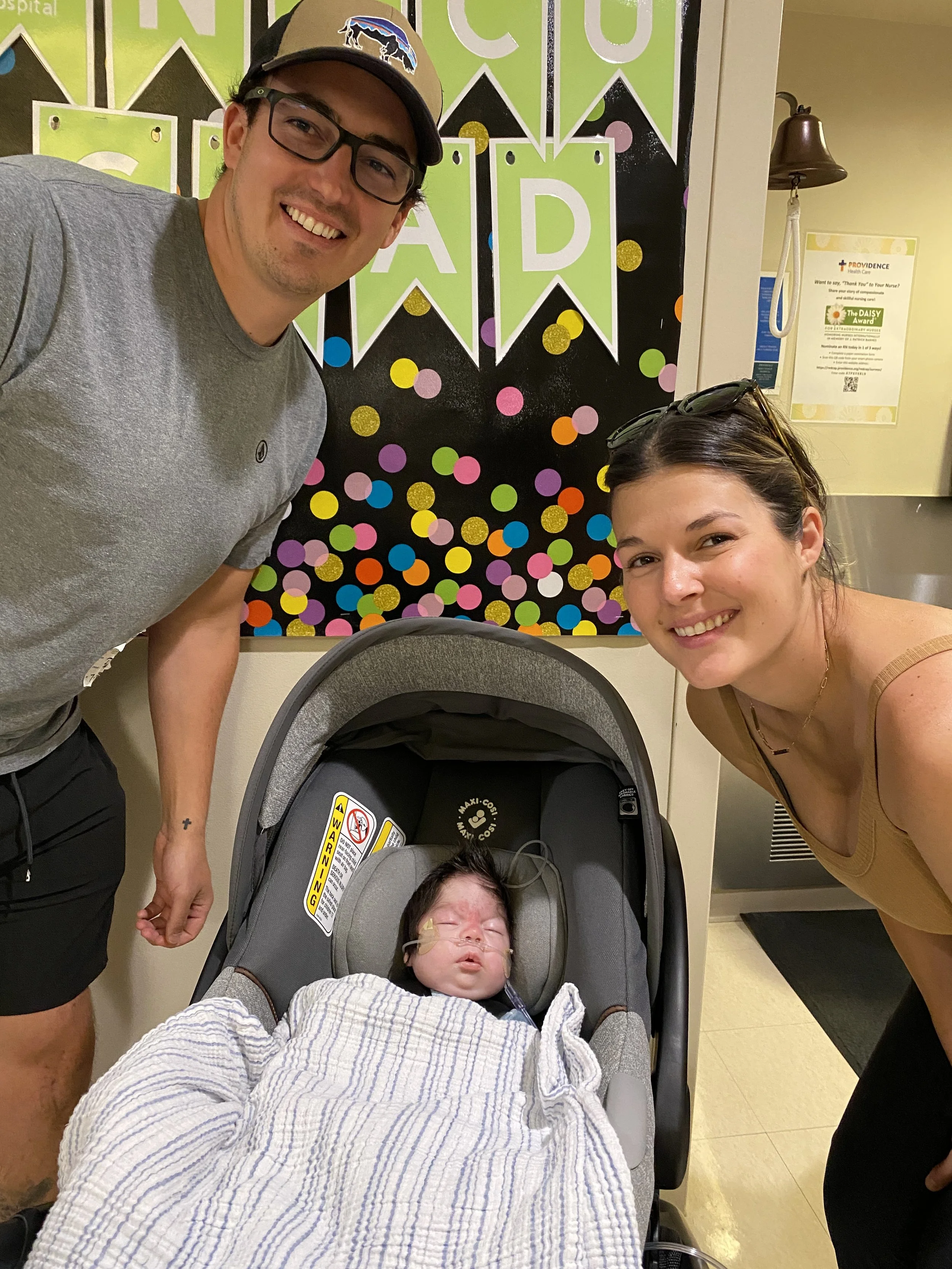 A smiling man wearing glasses and a cap with a buffalo logo, a woman with sunglasses on her head, and a baby sleeping in a car seat are gathered in front of a black background decorated with colorful dots and green banners with initials 'A D' at an hospital.