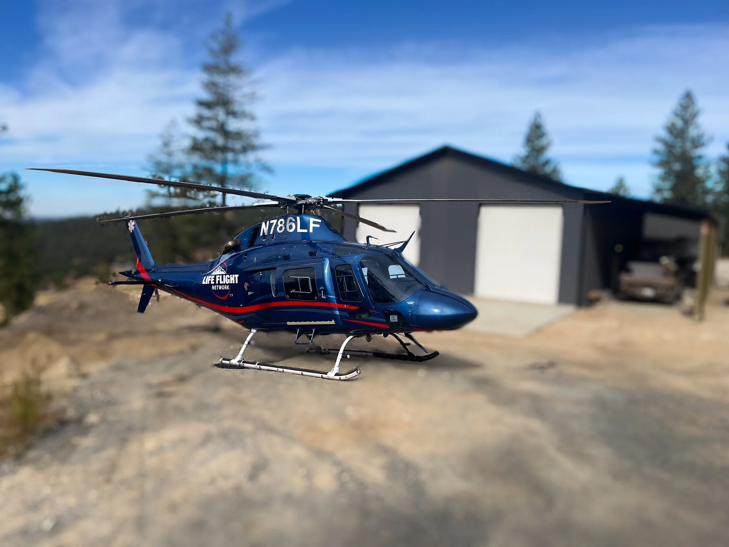 A remote-controlled helicopter model parked outdoors on a dirt surface, with a background of trees and a building under a blue sky.