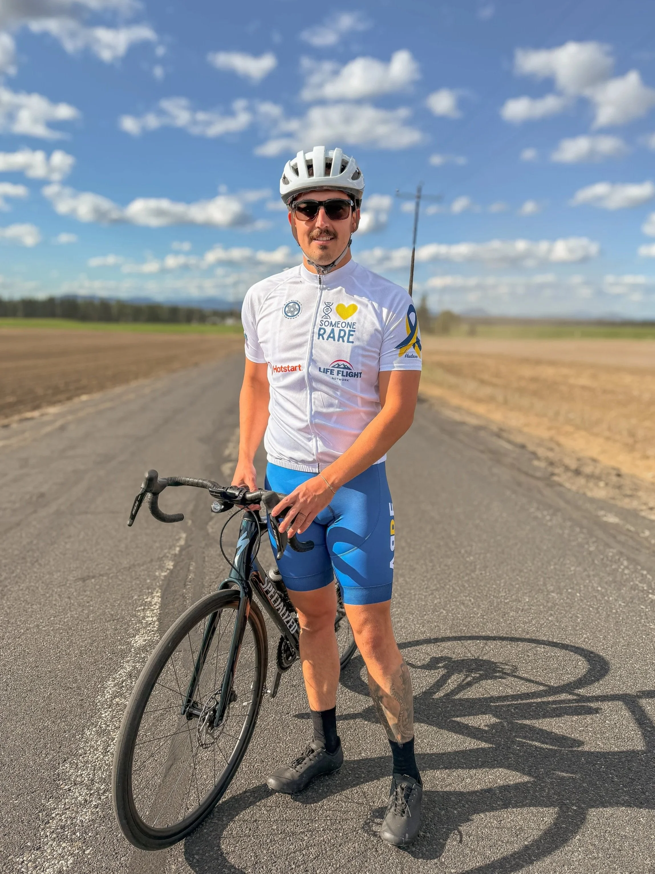 A man wearing cycling gear, including a helmet, sunglasses, and a white cycling jersey, stands with a black road bike on an open road. The background shows a clear blue sky with scattered clouds and a rural landscape.