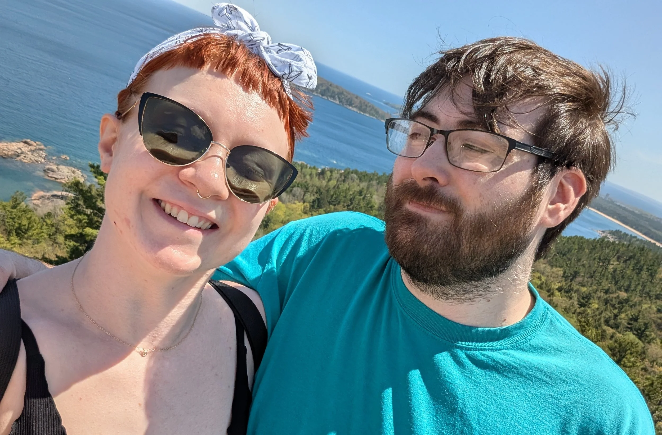 A woman with short red hair, sunglasses, and a bandana is smiling. A bearded man with glasses and brown hair is next to her. They are outdoors with a scenic ocean view, rocky coastline, and green trees in the background.