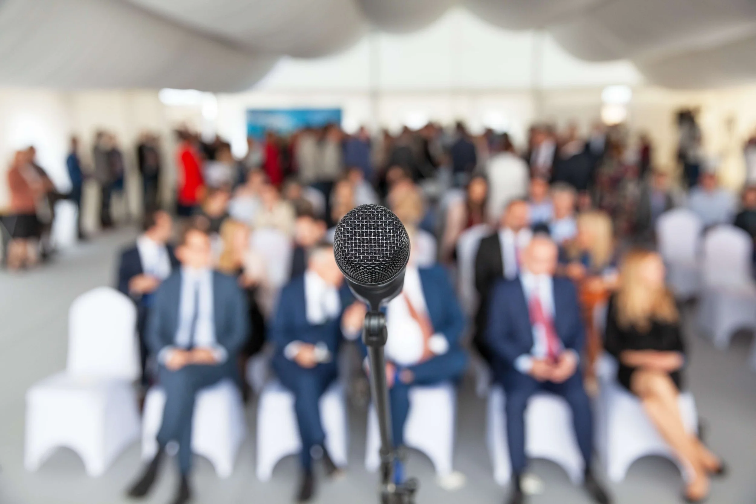 A microphone in the foreground with a blurred audience of people sitting and standing in a large room, suggesting a conference or presentation setting.