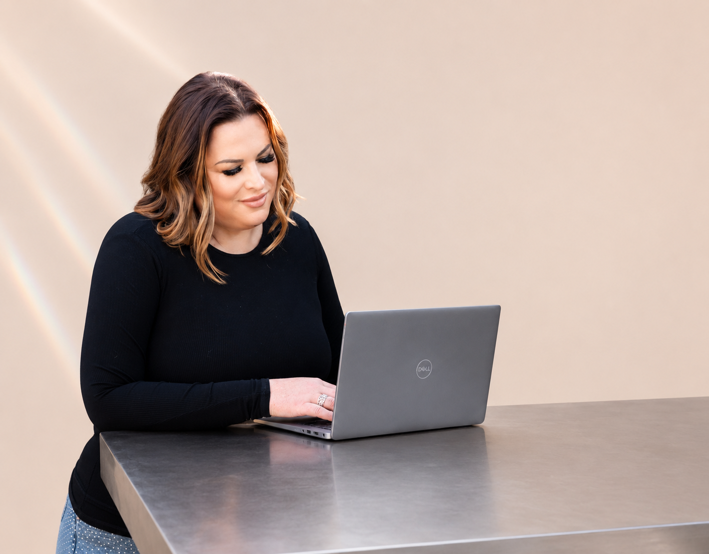 Leadership coach, keynote speaker, and author Alexandra Gerritsen is working on a silver Dell laptop at a gray table against a plain beige wall.